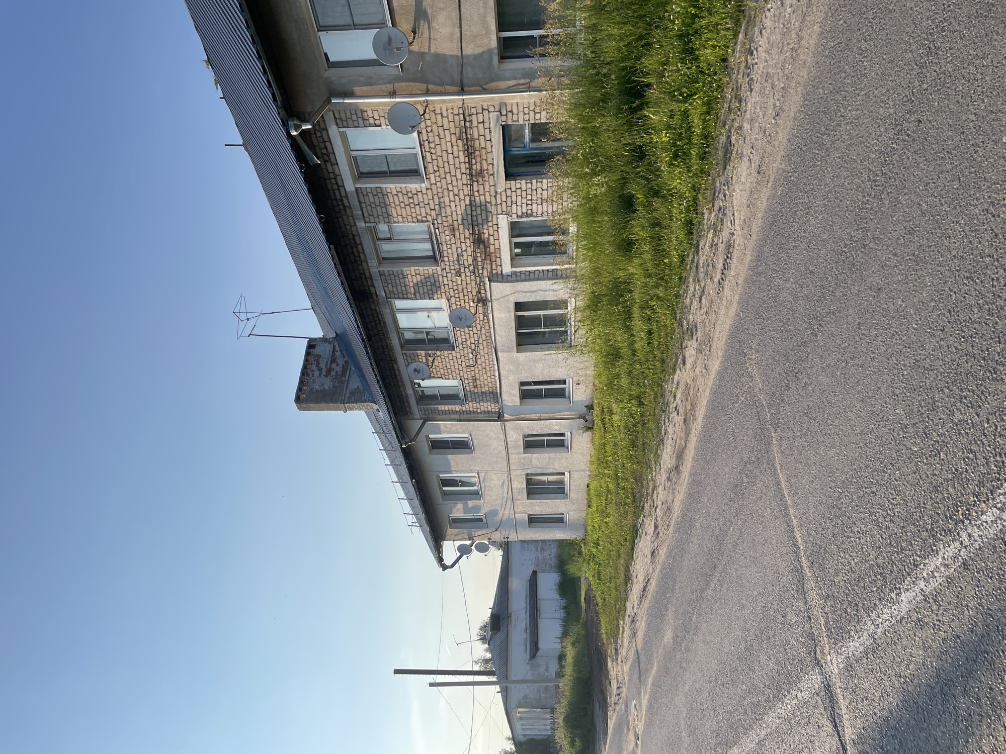 This is a photograph of a residential building in Пруды, Russia. The building appears to be multi-story with a brick facade on the upper floors and a lighter colored facade on the lower floors. It has numerous windows and several satellite dishes mounted on the exterior. A metal roof structure extends over part of the building.
Adjacent to the building is a strip of green grass and then a paved road with a white line marking. The sky is clear and blue, suggesting a sunny day. The image is taken from a low angle, looking up towards the building and sky. The sun appears to be low in the sky, casting shadows and illuminating the scene, indicating it is either early morning or late afternoon.
There are no people visible in the image. The scene depicts a quiet, everyday setting, likely a residential area. The overall impression is one of a typical day in a Russian town or village. There is no visible text in the image.