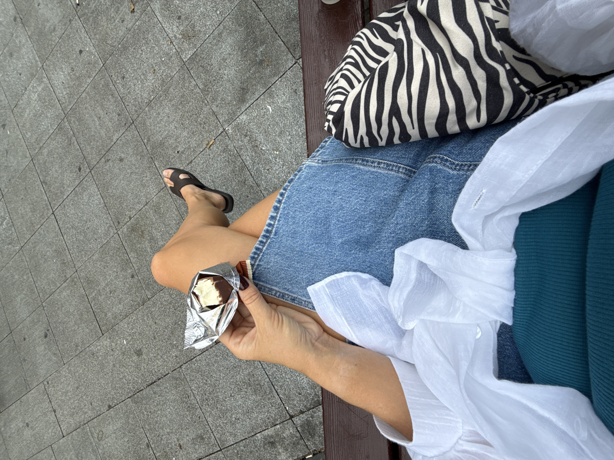 A person is sitting on a wooden bench outdoors, with their legs crossed. They are holding a partially unwrapped chocolate ice cream bar in their right hand, which is resting on their lap. The person is wearing a white linen shirt, a denim skirt, and dark sandals. Next to them on the bench is a bag with a zebra print. The ground in front of the bench is paved with square gray tiles.