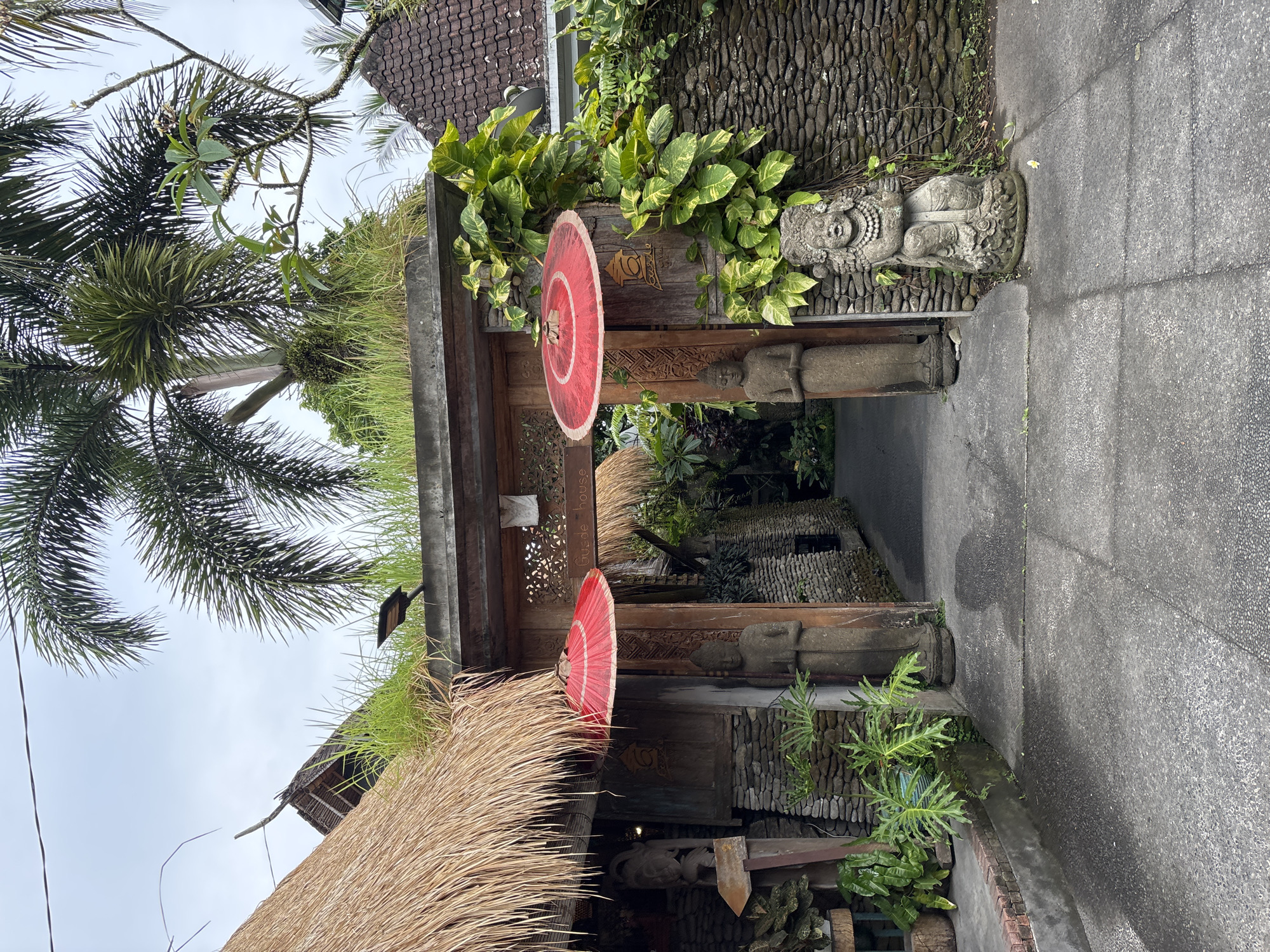 In Ubud District, Indonesia, a picturesque entryway to a property is depicted. The scene is dominated by a weathered wooden archway adorned with two vibrant red parasols. Flanking the archway are two stone statues, one on each side, contributing to the traditional Balinese aesthetic. To the right of the arch, a third, more ornate stone statue sits in front of a textured stone wall covered in lush green foliage.
The entrance itself leads to a pathway lined with stone walls and vegetation, suggesting a hidden garden or courtyard beyond. Above the archway, the roof of the structure is covered in thick thatch, blending seamlessly with the surrounding greenery that appears to grow on top of the building's upper levels. Tall palm trees with broad fronds extend into the cloudy sky, framing the scene and adding to the tropical ambiance.
The foreground consists of a textured, gray concrete pathway, wet in places, with visible seams and slight variations in color. The overall impression is one of tranquil beauty and a harmonious integration of nature and architecture.