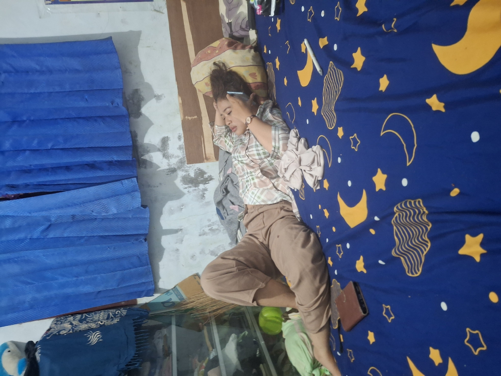 A young woman is lying on a bed with blue sheets decorated with yellow moons and stars. She is on her left side, propped up on her elbow and talking on a silver cell phone. She is wearing a plaid shirt and loose tan pants. To her left is a brown wallet and a light green object, possibly a pillow. Behind her is a wooden headboard, a pillow, and a stack of folded clothes. In the background, a blue curtain hangs above a shelf unit filled with various items, including a blue and white stuffed animal, a dark blue scarf with a patterned fringe, and a green ball. The wall behind the bed is textured and appears to be painted white. The setting appears to be a bedroom in Biak, Indonesia.