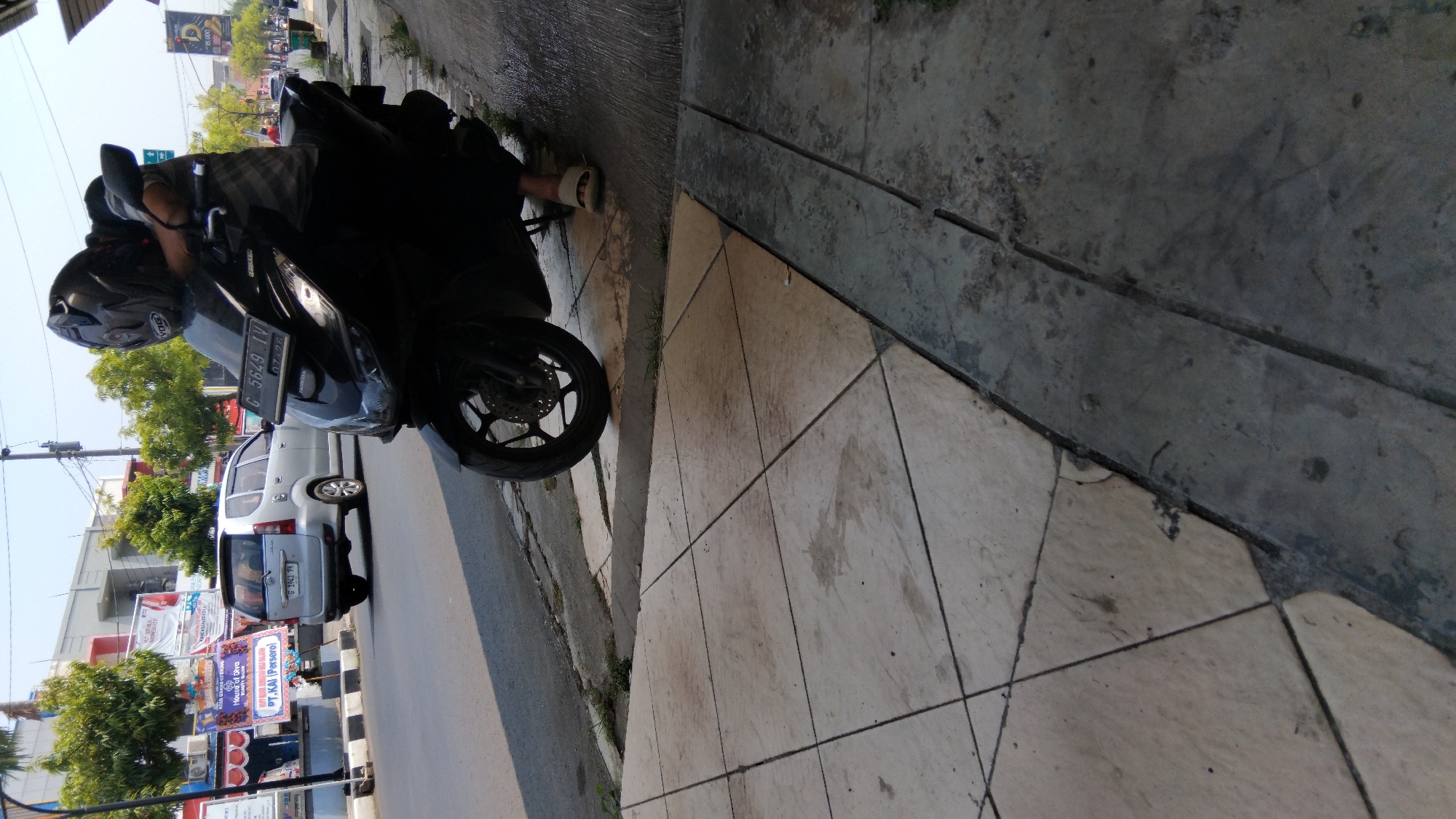 In Batang, Indonesia, a person wearing a helmet is leaning on a black motorcycle parked on a sidewalk. The motorcycle's license plate reads "G 5649 TV". The sidewalk is made of light-colored square tiles. Behind the motorcycle, a silver car is driving on the road, with advertisements and trees visible on the other side of the street. The sky is clear and blue.