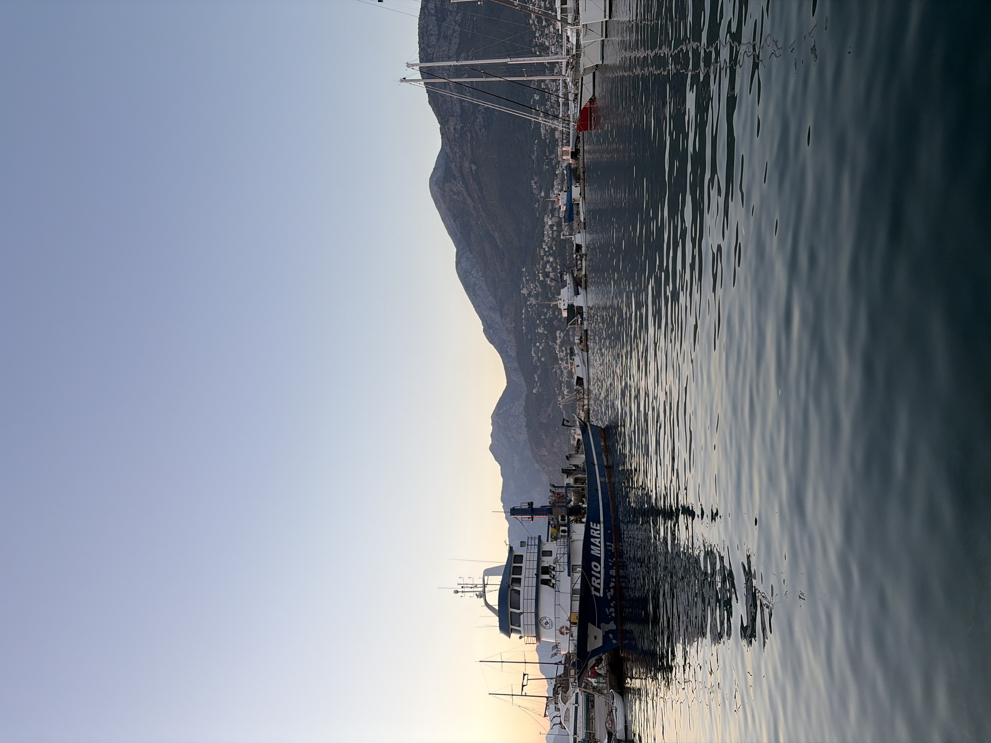 The image depicts a tranquil harbor scene in the city of Bar, Montenegro, likely at sunset or early morning given the soft, diffused light. The water is calm, reflecting the boats and the distant mountains. In the foreground, the dark, rippling water dominates the lower half of the frame.
A prominent blue and white fishing trawler, named "TRIO MARE," is docked on the left side of the image, taking up a significant portion of the mid-ground. It appears to be a substantial vessel with multiple decks and equipment visible. Smaller boats are moored alongside it and further in the background.
To the right of the trawler, several sailboats with tall masts are anchored, their white sails furled. One red-hulled sailboat is visible among them.
In the background, a range of dark, tree-covered mountains rises above the city. Small buildings and lights dot the mountainside, indicating a populated area. The sky above is a clear, pale blue, transitioning to a lighter shade near the horizon where a faint glow suggests the sun is low. The overall atmosphere is peaceful and serene. No people are discernible in the image.