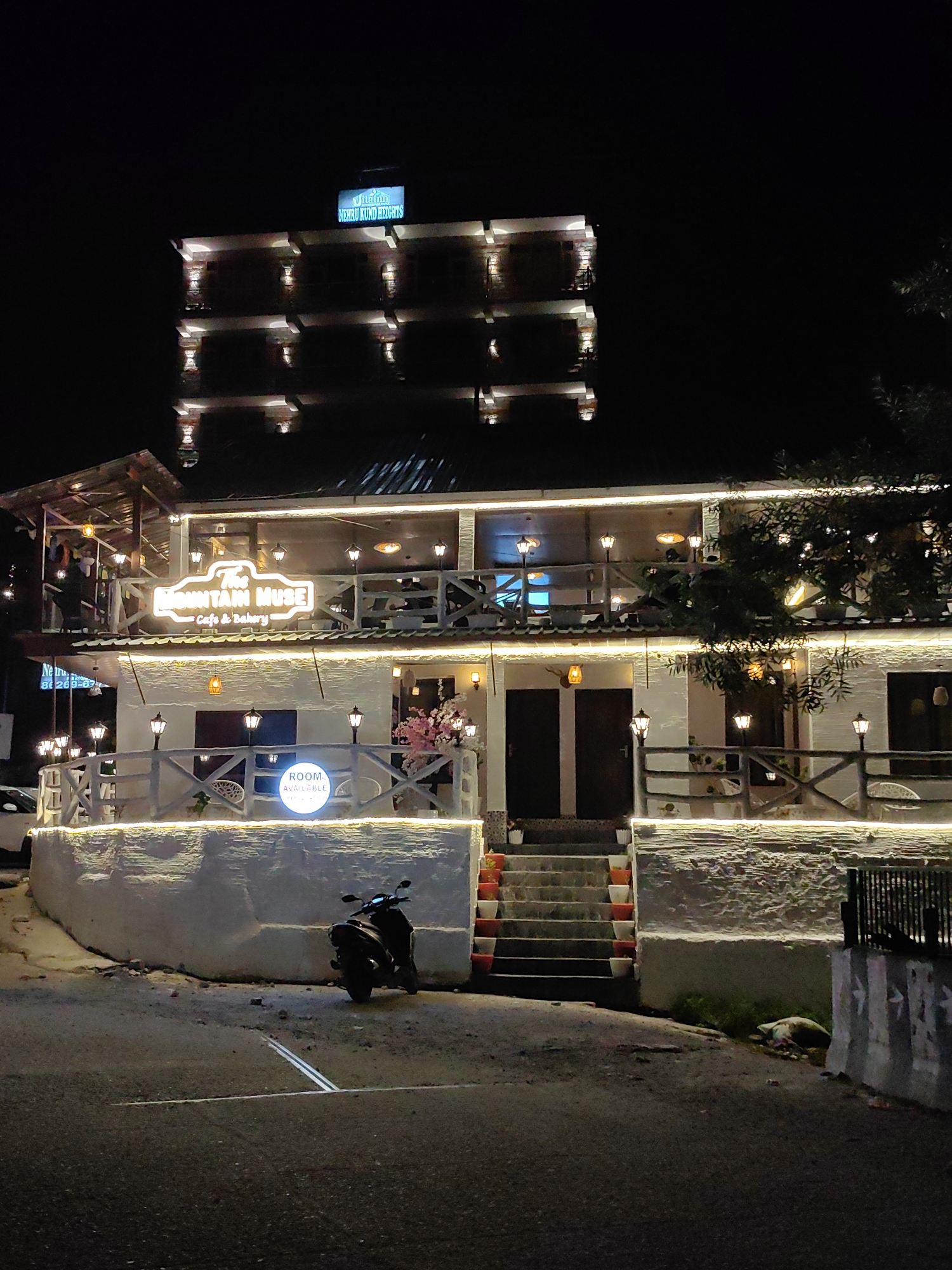 The image shows "The Mountain Muse Cafe & Bakery" at night. The building is illuminated by numerous string lights and decorative lamps. Above the cafe, a taller building with the sign "Nehru Kund Heights" is visible. A scooter is parked on the left side of the road in front of the cafe. There are stairs leading up to the cafe entrance, decorated with potted plants. The cafe has a wooden railing and a sign that says "ROOM AVAILABLE". The setting appears to be a street in Barheta, India.