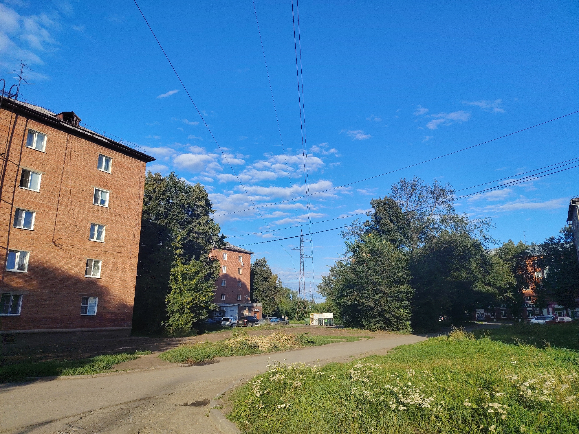 This is a wide-angle, ground-level, outdoor photograph of a street scene in Kemerovo, Russia, on a clear, sunny day with scattered clouds. The image is dominated by a vibrant blue sky, traversed by several high-voltage power lines.
In the left foreground, a multi-story brick apartment building, with red bricks and white-framed windows, stands partially in shadow. Several windows are open, and some have external wiring. An antenna is visible on the roof. Adjacent to the building, a paved road curves towards the right side of the frame, bordered by a grassy verge with white wildflowers and green grass.
In the middle ground, the road opens into a dirt area where several parked vehicles, including cars and vans, are visible. Beyond the vehicles, a tall metal electricity pylon stands prominently against the sky, with numerous power lines originating from it. A small, utilitarian-looking white structure, possibly a kiosk or utility shed, is situated near the pylon.
The right side of the mid-ground is filled with dense green trees. Further back, another brick building, appearing to be a residential or commercial establishment, is partially visible through the foliage. More trees and buildings are visible in the distance, suggesting an urban or suburban environment. A few individuals are casually standing near the vehicles in the mid-ground. The overall impression is one of a typical residential area with supporting infrastructure.