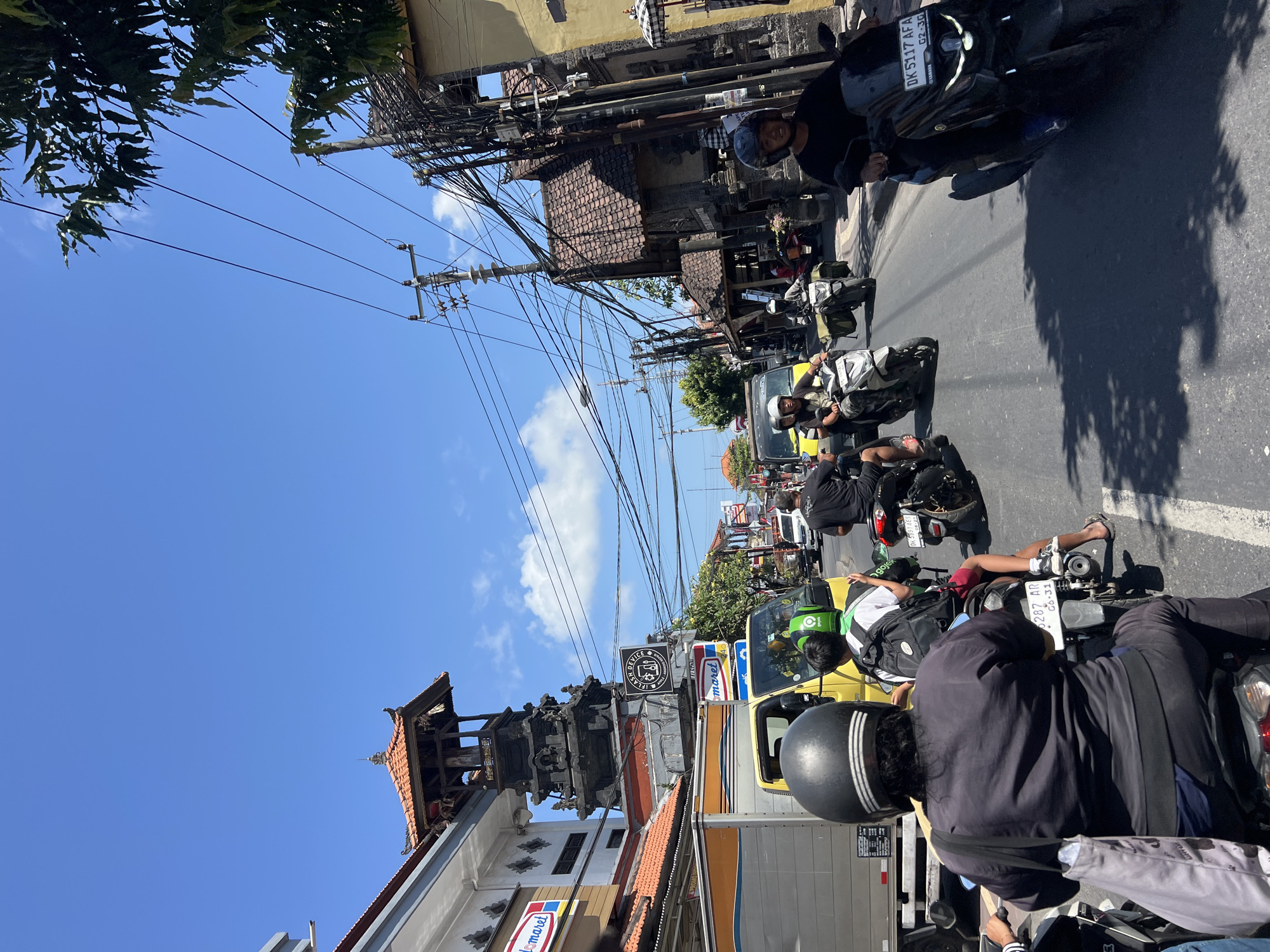 The image depicts a busy street scene in Denpasar, Indonesia, characterized by heavy motorcycle traffic under a clear blue sky. In the foreground, a person wearing a black helmet and dark clothing rides a motorcycle, with a grey tote bag slung over their shoulder. Behind them, another motorcycle carries a rider in a green helmet and a passenger with a backpack. A yellow truck is visible in the middle ground, alongside other motorcycles and cars.
The architecture of the buildings lining the street suggests a traditional Indonesian style, with ornate tiled roofs and intricate detailing, particularly noticeable on a multi-tiered structure to the left of the frame. Electrical wires are prominently strung across the street, a common sight in many urban areas in Indonesia. Signs for "Indomaret," a convenience store chain, are visible on buildings, indicating commercial activity. The overall atmosphere is one of a lively, bustling urban environment on a sunny day.