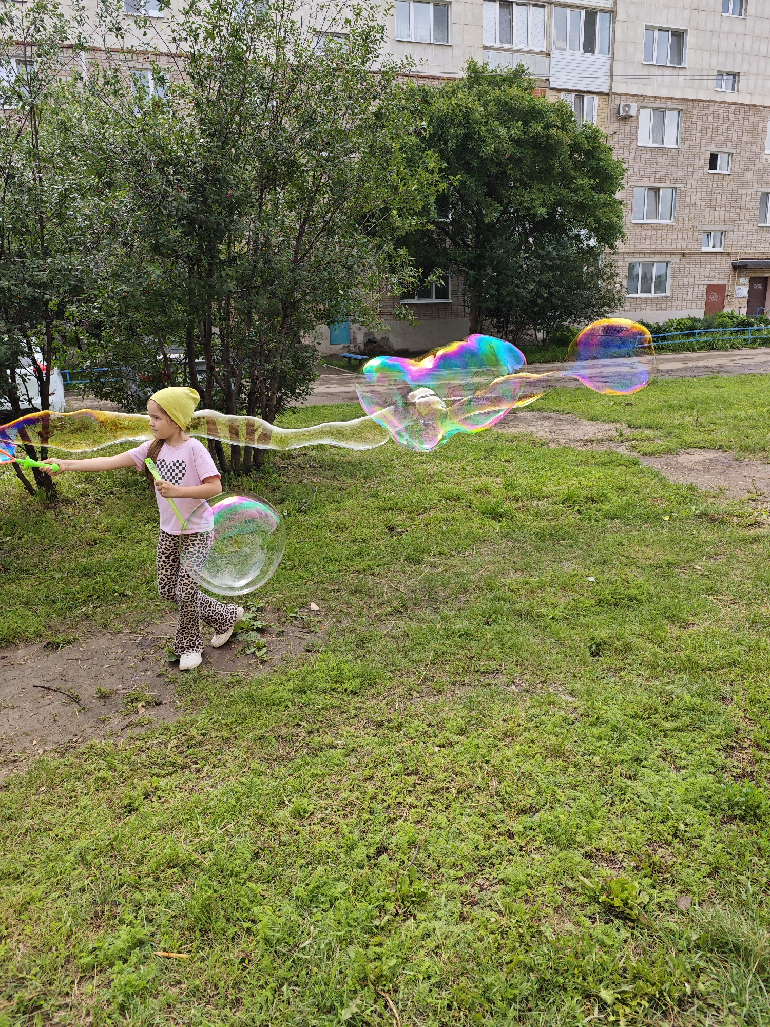 In the city of Tujmazy, Russia, a young girl wearing a yellow hat and a pink t-shirt with a black checkered heart is blowing giant, iridescent soap bubbles in a grassy area. She is holding a green wand with a checkerboard pattern, extending it to her right as a long trail of bubbles flows away from her. One large, perfectly spherical bubble floats near her feet, while a chain of connected bubbles, displaying a rainbow of colors, stretches horizontally across the scene. Behind the girl, a building with light-colored brickwork and multiple windows rises, flanked by lush green trees. The ground is a mix of dirt paths and green grass, indicating an outdoor, possibly residential, setting.