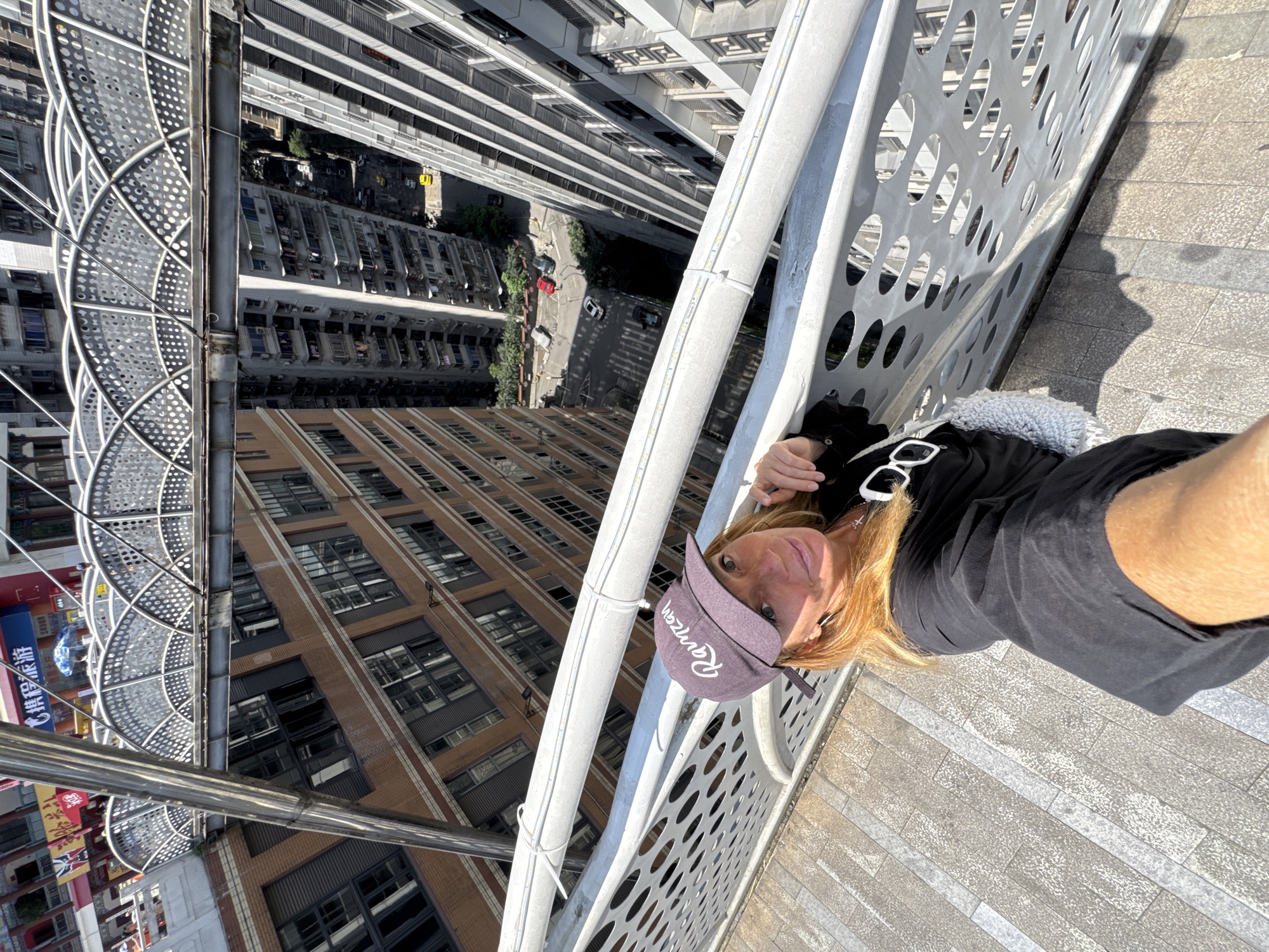 A woman is taking a selfie on the rooftop of a building in Yuzhong District, China. The backdrop includes high-rise buildings and a spiral staircase. The woman is wearing a black shirt and a lavender cap with "Ranzan" written on it. Sunglasses are placed on her shirt near the shoulder. The environment is urban with visible streets and traffic. The photograph is taken from a high angle, looking down into the city. The woman appears to be interacting with the camera, suggesting she is taking the photo herself.