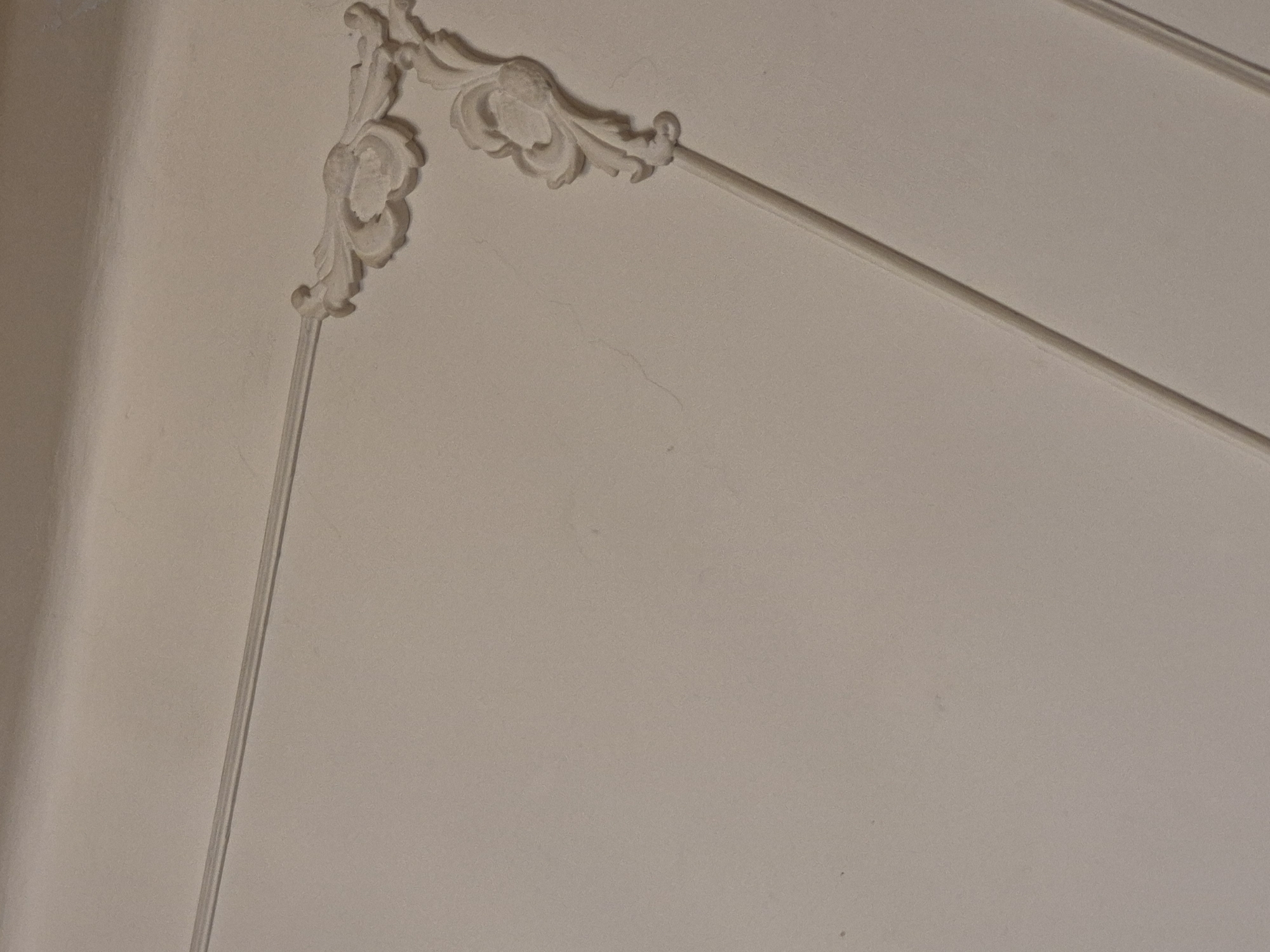The image displays a close-up view of ornate white ceiling molding against a plain, light-colored wall. The molding features decorative floral elements and geometric lines, forming a corner. The lighting suggests an indoor setting, possibly a room with a textured ceiling. The specific location in Yerevan, Armenia, cannot be determined from the visual information provided. There are no people or actions depicted in the image. The environment appears to be a static interior architectural detail.