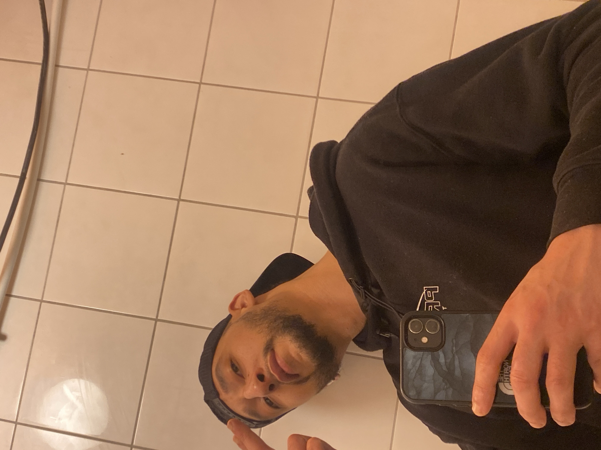 A male subject is depicted in a close-up, angled shot, leaning against a tiled wall in an indoor location within the city of Schiltigheim, France. He possesses light brown skin, a trimmed beard, and a mustache, and wears a dark baseball cap backward and a black hooded sweatshirt. His left index finger is extended towards his left temple. He holds a dark-colored smartphone with a blue-grey patterned case in his right hand, positioned in the lower-center of the frame, indicating he is taking a mirror selfie. The phone case has a visible "The North Face" sticker, which appears reversed due to reflection. The background consists of light-colored, square ceramic tiles with dark grout lines. A dark, thin cable or pipe runs horizontally across the upper portion of the tiled wall. The lighting suggests an artificial indoor environment.