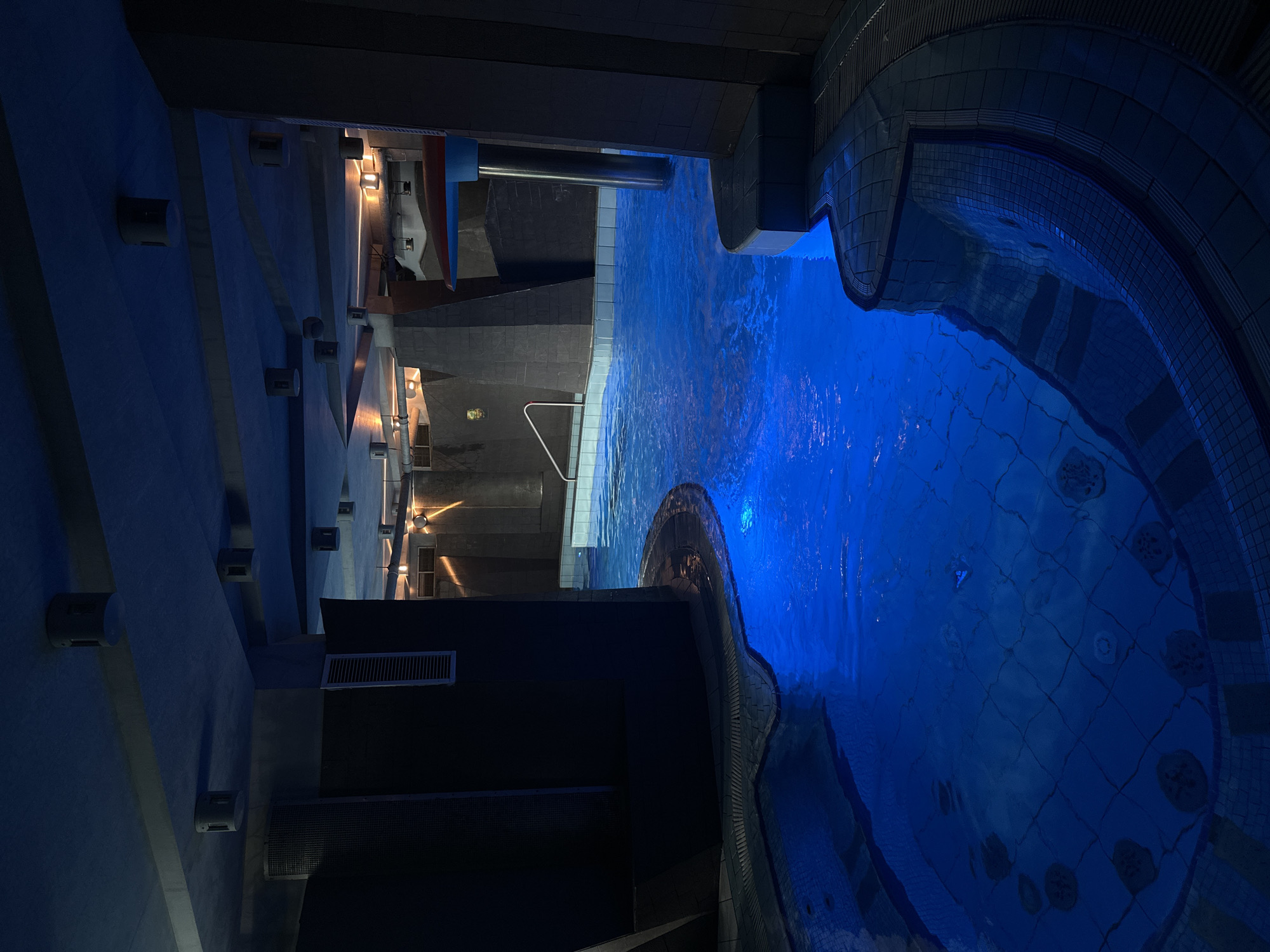 A dimly lit indoor swimming pool area is depicted, featuring bright blue illuminated water. The architecture appears modern and somewhat industrial, with concrete ceilings and angular structures. Multiple round, cylindrical light fixtures are visible on the ceiling. The pool itself has tiled edges and a slightly stepped design in the foreground. The overall atmosphere is serene and tranquil, suggesting a spa or wellness center. The image was taken in Minsk, Belarus.