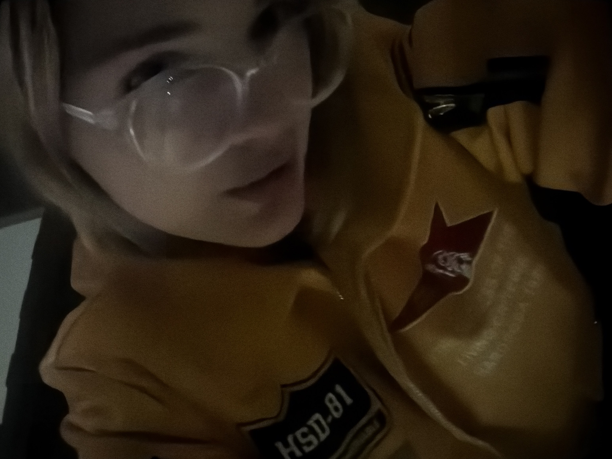 A person wearing glasses and a yellow jacket is visible in this low-lit, blurry image. The jacket has a patch with "HSD-81" on it and a red star design. A black object, possibly a bag strap or other accessory, is also present. It is not possible to determine the location of the image with the information available. It is also impossible to confirm it is located in Naberezhnye Chelny, Russia.