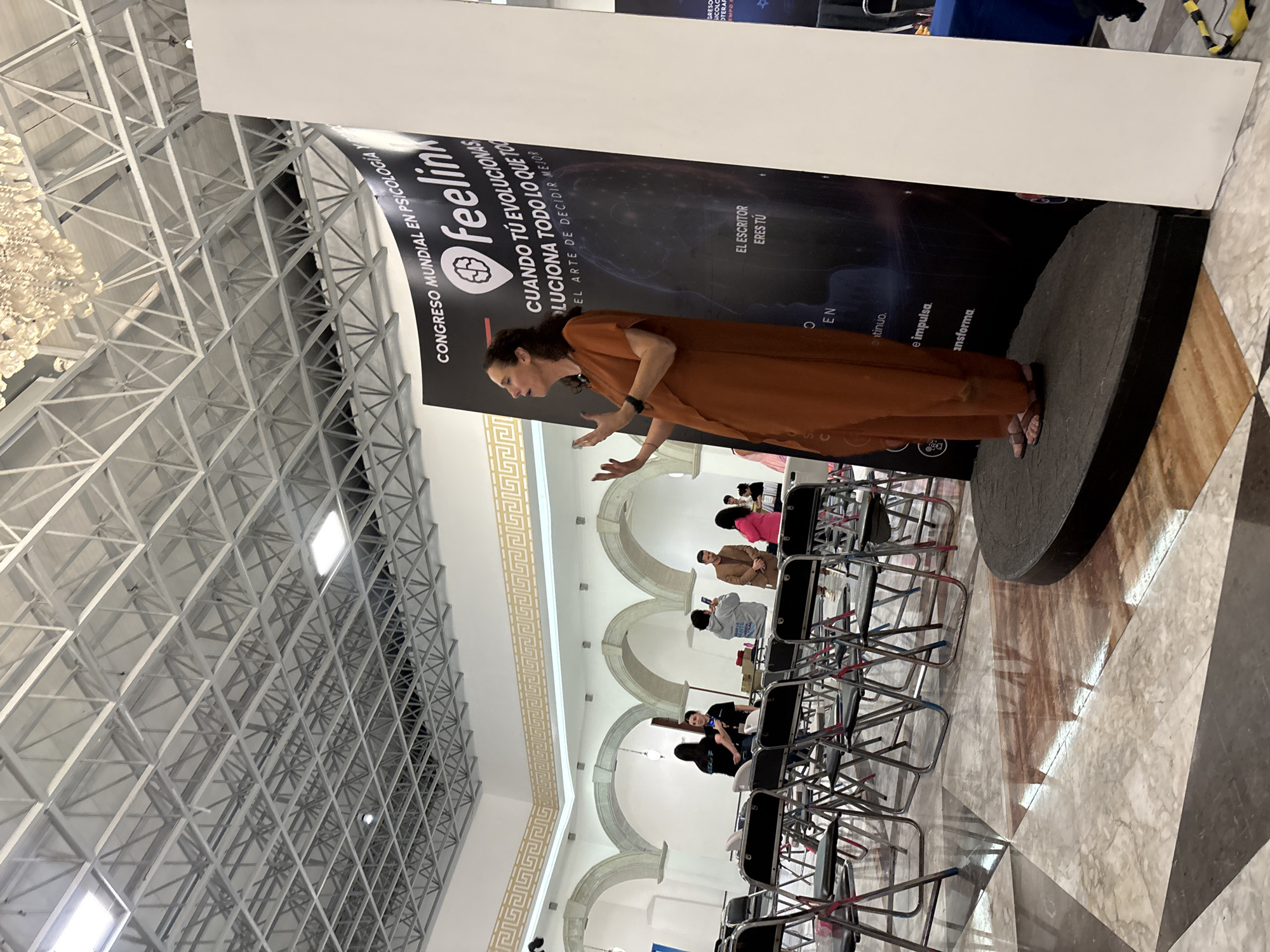 The photo shows a woman in a brown dress giving a presentation on a stage in a large room with a high ceiling. A banner behind her says "CONGRESO MUNDIAL EN PSICOLOGIA Feelin CUANDO TU EVOLUCIONAS LUCIONA TODO LO QUE TOU EL ARTE DE DECIDIR MEJOR EL ESCRITOR ERES TU". There are chairs set up in front of the stage for an audience. People can be seen in the background. The location is likely Mexico City, Mexico.