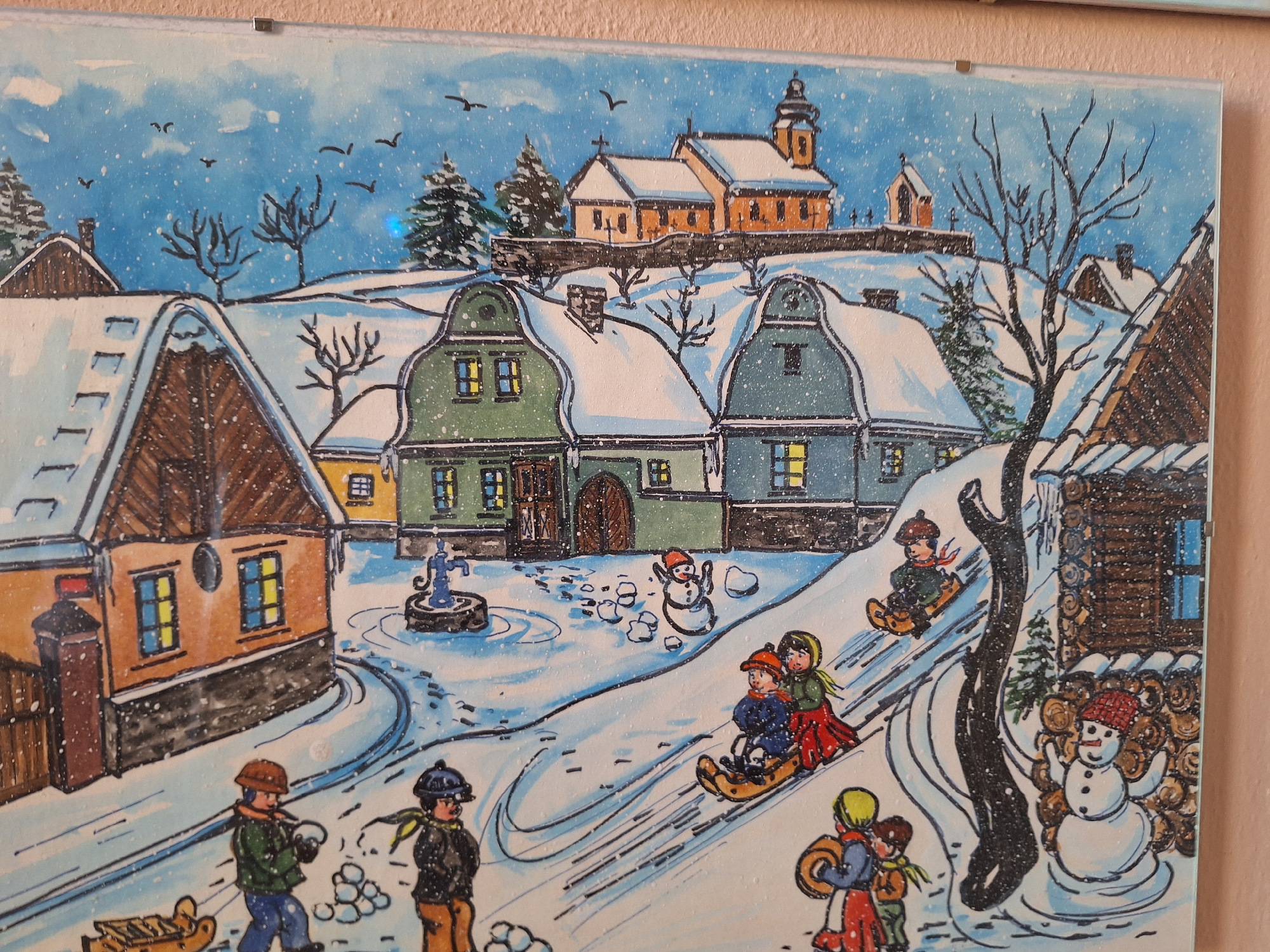 The image depicts a charming watercolor painting of a snowy winter village scene. The architecture suggests a European setting, with quaint, multi-story buildings with steeply pitched roofs covered in snow. Some buildings have visible chimneys emitting faint smoke, and windows glow with warm light, indicating occupancy. In the background, a church with a bell tower stands on a hill, typical of historical European towns.
The foreground is bustling with activity. Children are engaged in various winter pastimes. Several children are sledding down a snow-covered hill, one group on a single sled, with one child steering. Another child is standing near a snowman, which has a red hat and outstretched arms. In the lower left corner, two children are pulling a wooden sled, with one of them holding a snowball, suggesting play. Further left, another child is pulling a sled loaded with what appears to be firewood. A water pump stands in the middle of the scene, surrounded by snow-covered ground and footprints.
The sky is a bright blue, with scattered white clouds and falling snowflakes, creating a serene winter atmosphere. Several birds are depicted in flight against the sky. The overall style is naive and folksy, characteristic of traditional folk art. While the image captures a generic winter village, its style and architectural elements are reminiscent of depictions of historical towns in regions like Bohemia, which includes Prague in the Czech Republic.