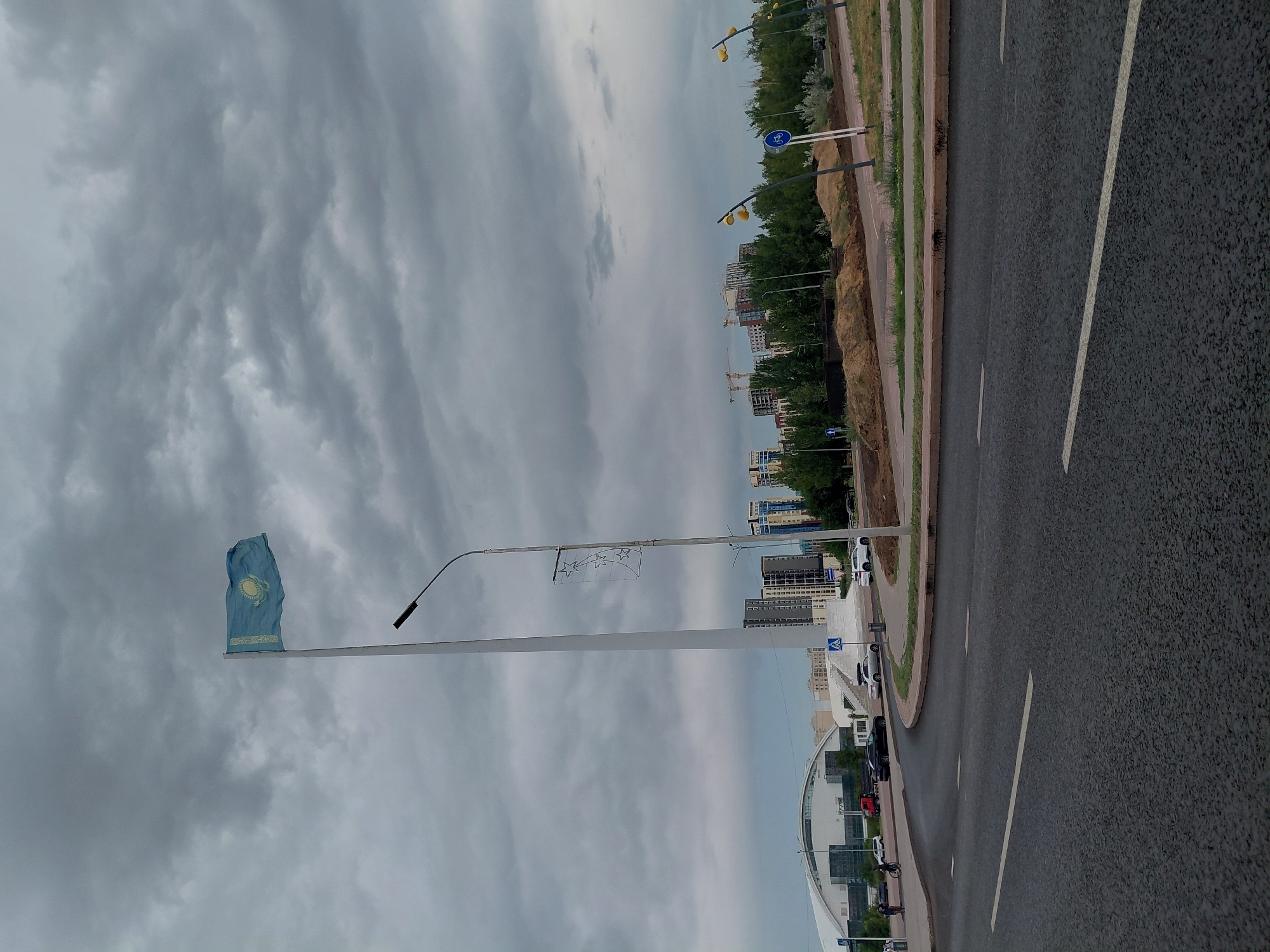 A wide shot captures the Kazakhstan flag flying high on a tall pole against a cloudy sky. In the background, modern buildings and construction cranes dot the skyline. A street curves in the foreground, with cars driving on it. The scene appears to be a public space in the city of Qarağandı, Kazakhstan, possibly near the city administration building, given the prominent flag and urban architecture.