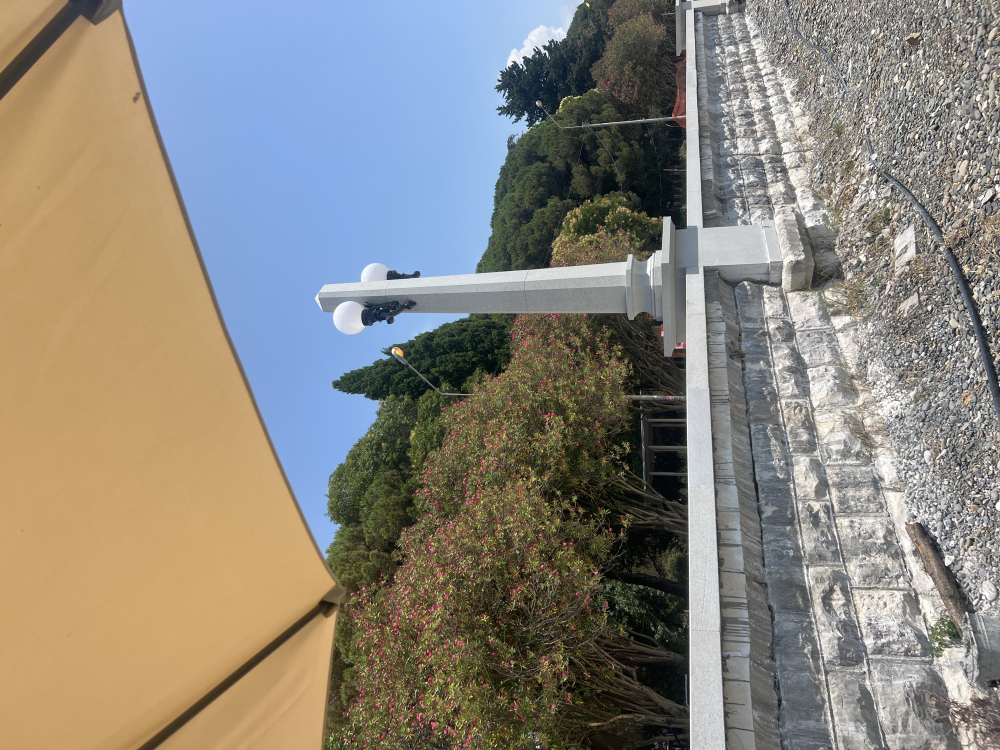 This image captures a section of the Sokhumi embankment in Abkhazia on a sunny day. A large, tan umbrella dominates the upper left portion of the frame, casting a shadow and suggesting an outdoor seating or relaxation area. In the mid-ground, a gray stone wall lined with what appears to be flowering trees with pink blossoms forms a prominent barrier. Behind the wall, a dense line of green trees, including cypress trees, creates a lush backdrop.
A tall, octagonal gray stone lamppost with two globe-shaped lights stands centrally on the wall. The wall itself is constructed of large, rough-hewn gray stones, with a smoother, lighter-colored stone cap. The ground in the foreground is covered with small, gray pebbles. A black cable runs across the pebbled area. The sky is a clear, bright blue. There are no people visible in the image, and the scene appears tranquil and well-maintained, consistent with a public promenade.