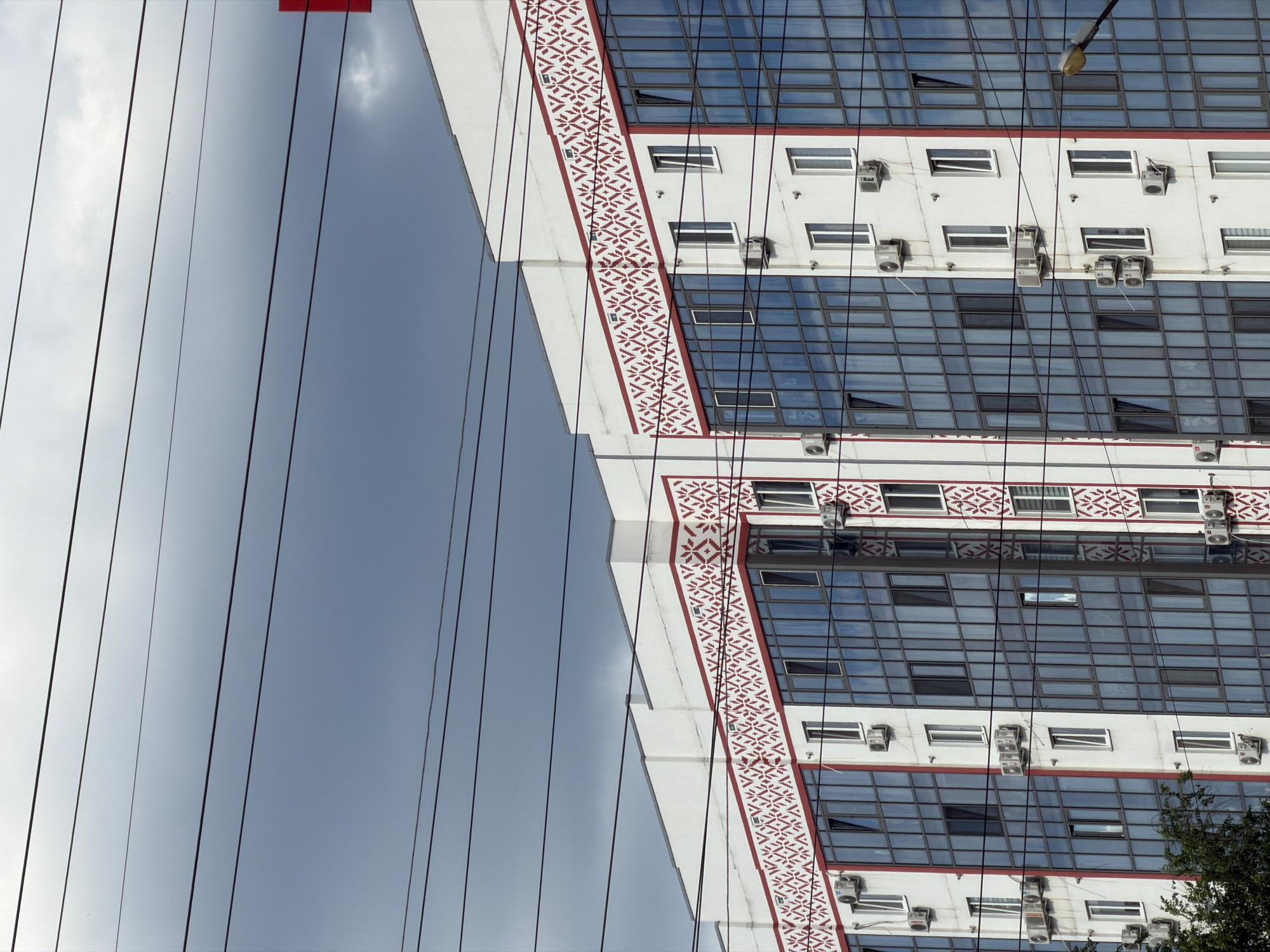 A close-up shot of a modern apartment building with a dark, cloudy sky as a backdrop. The building features a white facade with red decorative trim above a series of large, blue-tinted windows. Several air conditioning units are visible mounted on the exterior walls. Multiple power lines crisscross the frame, obscuring parts of the sky and building. The bottom of the image shows a glimpse of green foliage from trees. No people or vehicles are visible.
