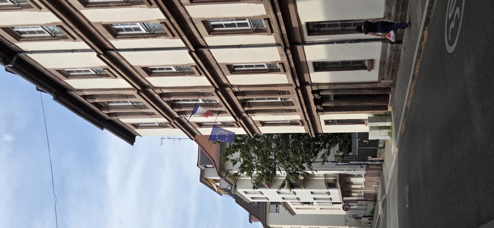 The image depicts a street scene in Strasbourg, France. A woman, wearing a dark patterned top and dark pants, walks away from the camera on a sidewalk. She carries a red and white plastic bag in her right hand. She is walking past a light-colored building with brown trim and multiple windows. Two flags are displayed on the side of this building: a French flag and a European Union flag. To the left of this building, other buildings are visible, including a white multi-story building with balconies and a sloped roofline. The street is paved with asphalt, and a speed limit sign (30) is visible on the road. Trees are present along the sidewalk, and the sky is partly cloudy. The architecture suggests an urban environment, possibly a residential or administrative area.