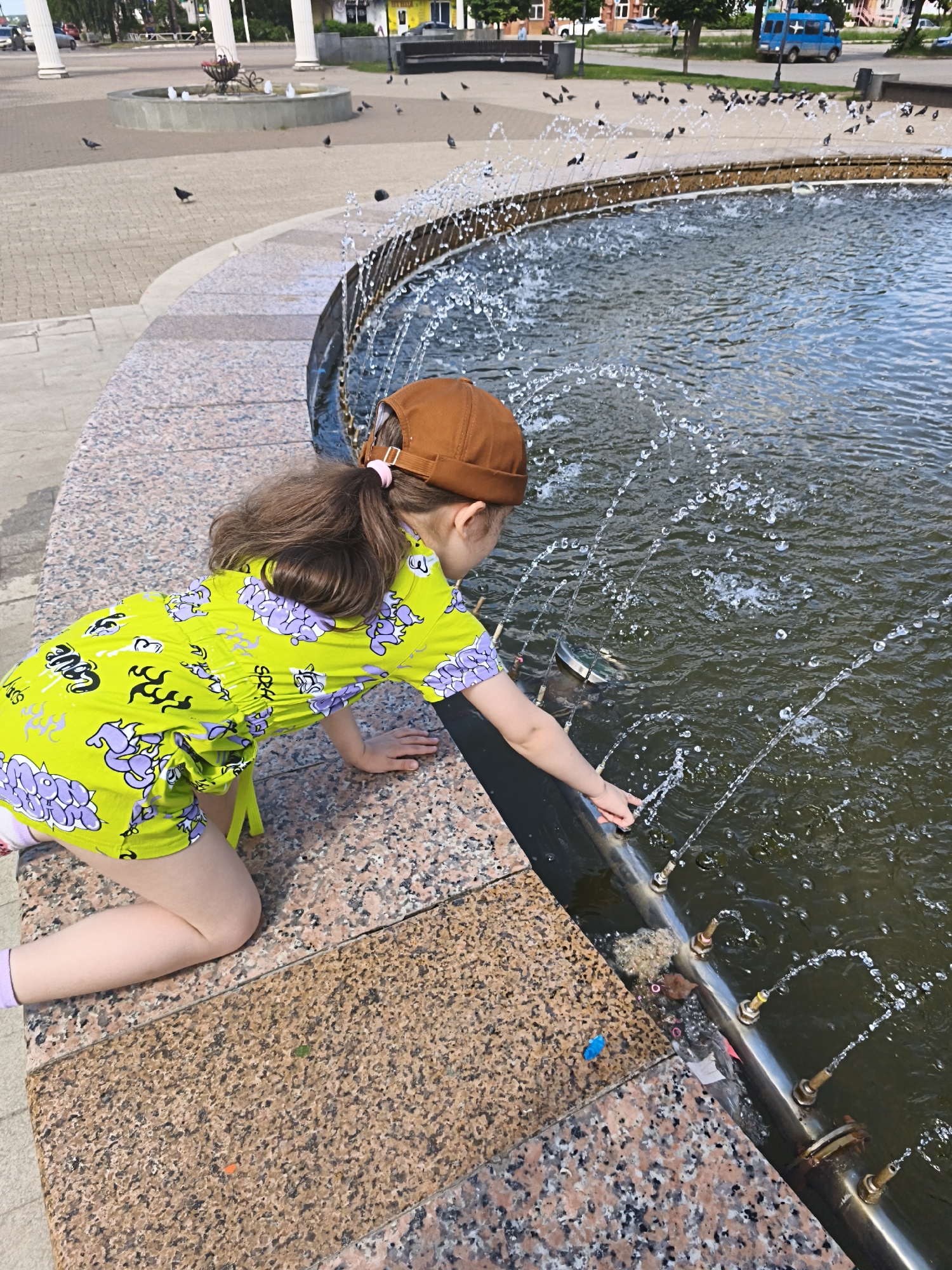 A young individual, identified as a child, is depicted in a daytime outdoor setting in what appears to be a public square in the city of Chaikovsky, Russia. The child, wearing a brown baseball cap and a vibrant lime green two-piece outfit adorned with purple and black graffiti-style patterns, is kneeling on a speckled granite perimeter of a large, circular fountain. Their dark hair is pulled back in a ponytail secured with a pink elastic. The child's right hand is extended into the fountain, with fingers touching or approaching the water jets emanating from a metallic pipe structure at the water's surface.
The fountain's water is dark and agitated by multiple streams spraying upwards and inwards. Debris, including a blue plastic object and general sediment, is visible within the fountain's water near the edge.
In the mid-ground and background, a paved pedestrian area with light-colored pavers extends. Numerous pigeons are scattered across this area, with some in flight and others foraging on the ground. To the left, a smaller, tiered fountain and several white architectural columns are visible. Vehicles, including a blue van, are present on a street in the background, bordered by buildings and trees. The scene is illuminated by bright daylight, casting short shadows.