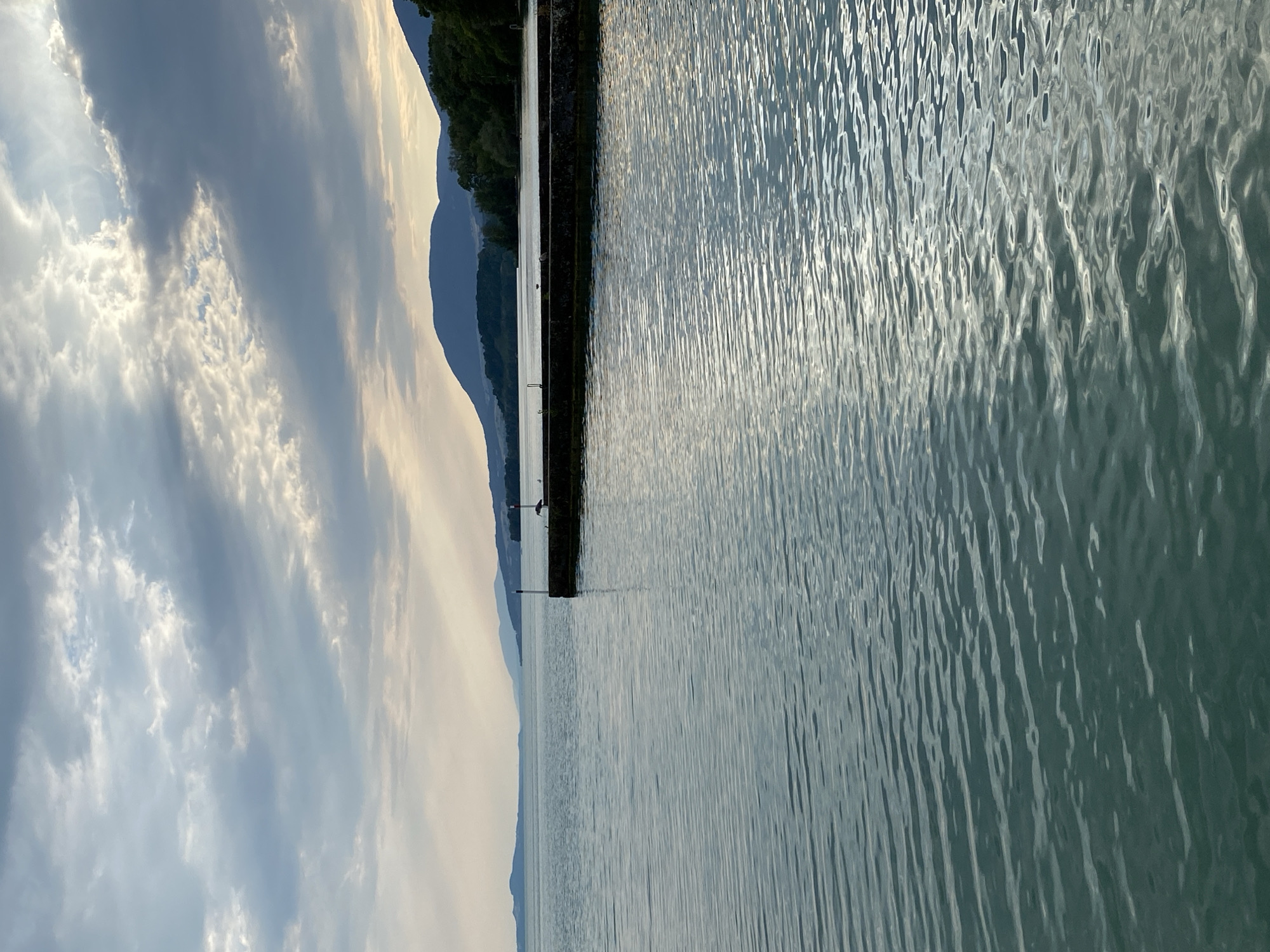 The image depicts a serene lakeside scene at Lake Neuchâtel in Cortaillod, Switzerland. The foreground is dominated by calm, rippling water, reflecting the muted tones of the sky. In the mid-ground, a stone pier extends into the lake, with two white posts and a red beacon visible at its end. A bird, possibly a cormorant or similar waterbird, stands on the pier, facing away from the camera. In the background, rolling hills covered in dark green trees form a natural landscape that meets the horizon. The sky is characterized by scattered clouds, with shafts of light breaking through, suggesting either early morning or late afternoon light. The overall atmosphere is peaceful and tranquil.