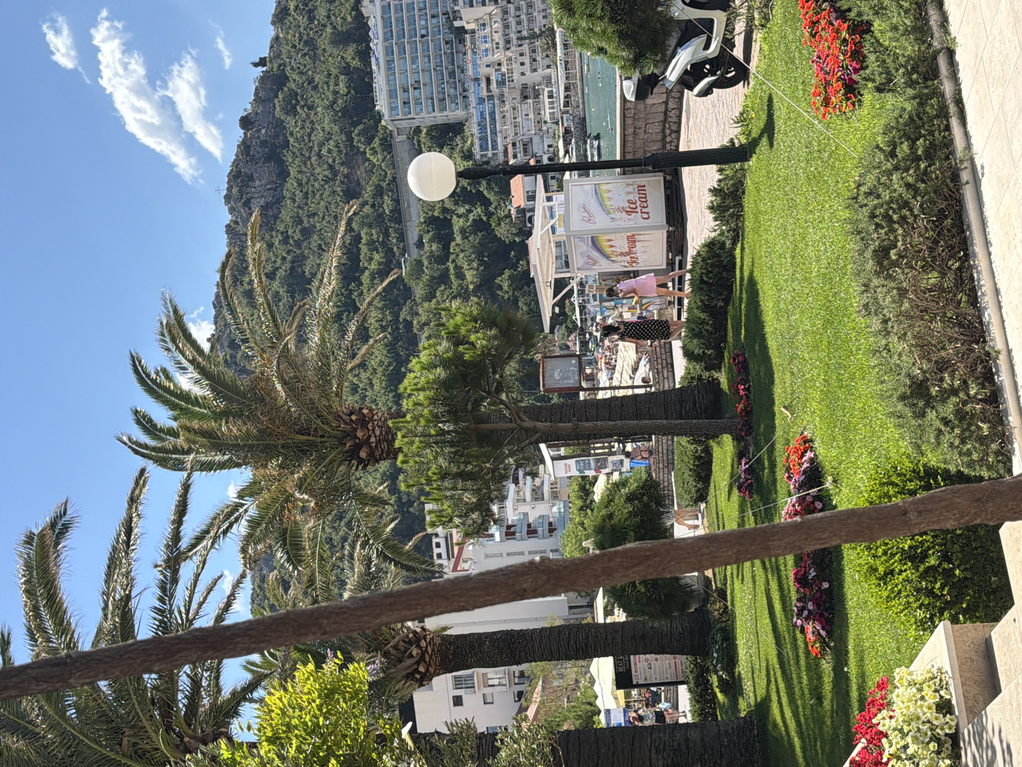 This media file depicts an outdoor scene with a focus on a lush green lawn bordered by flower beds. In the foreground, a thick rope stretches across the frame, suggesting it might be part of a barrier or railing. Several palm trees are visible, their fronds reaching towards a bright blue sky dotted with white, wispy clouds.
In the mid-ground, there is a paved walkway with a lamppost. Behind the walkway, a cluster of buildings rises up, suggesting a town or resort area nestled against a green, mountainous backdrop. Some of these buildings appear to be hotels or apartment complexes. Further in the distance, the sea or a large body of water can be seen.
On the walkway, several people are visible. Two individuals, one in a pink dress and another in a polka-dot dress, appear to be walking. There are also other figures further back, suggesting a public area with some activity. One stall or kiosk is visible with signage that includes the words "Ice cream."
The scene is bathed in bright sunlight, casting distinct shadows from the palm trees and lamppost onto the grass, indicating it is likely daytime, possibly late morning or early afternoon. The overall atmosphere appears relaxed and pleasant, consistent with a vacation or leisure setting. The presence of palm trees, the clear blue sky, and the architecture hint at a warm, possibly Mediterranean or coastal location.