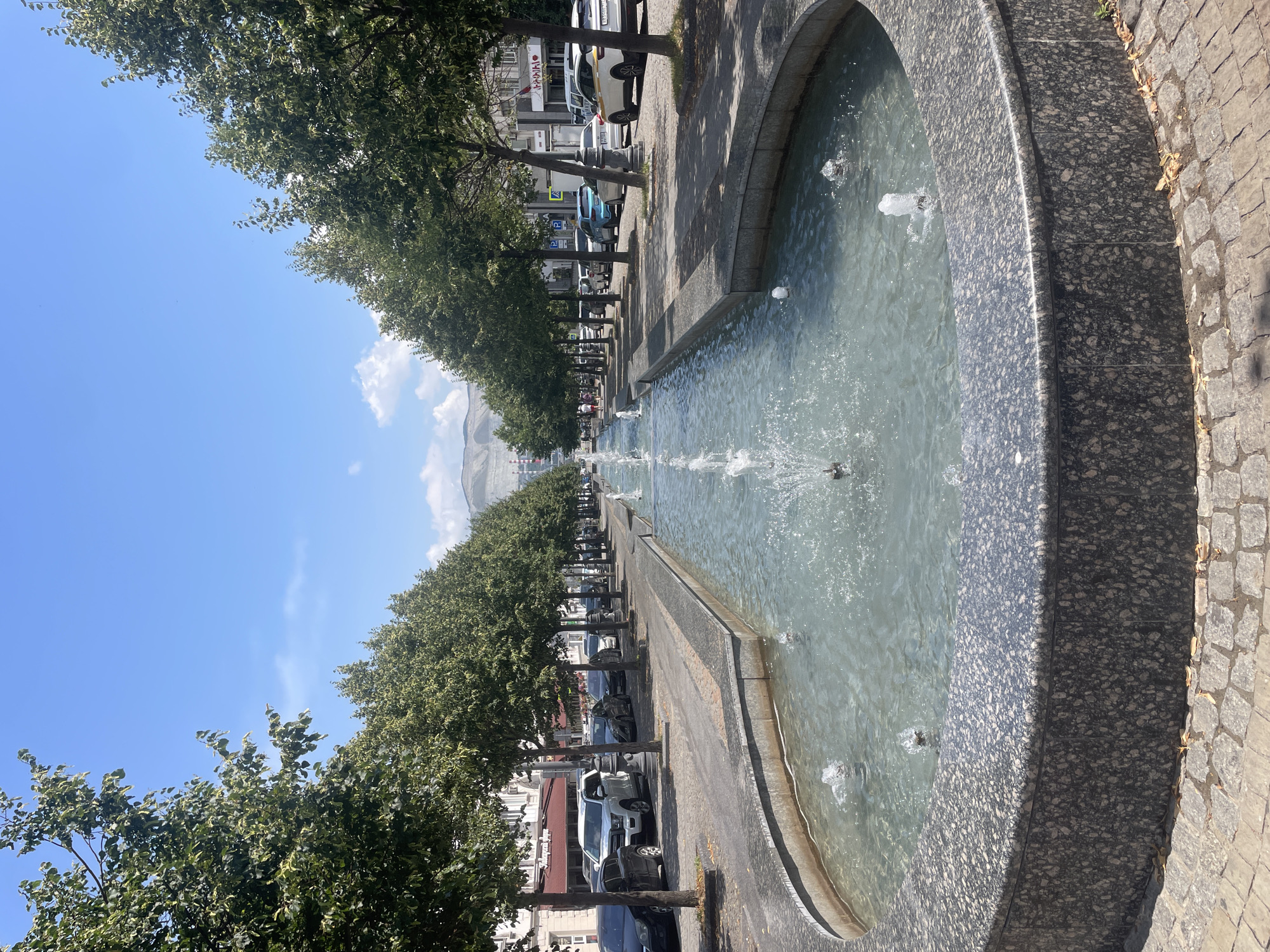 The image depicts a long, narrow fountain with multiple spouts of water in motion, situated in a city plaza or park. The fountain is lined with granite and is surrounded by a stone walkway. On either side of the fountain, a wide sidewalk is lined with numerous trees, casting shadows on the ground. Beyond the trees, parked cars are visible along the street, with buildings in the background. The sky is clear blue with a few wispy clouds. In the distance, mountains can be seen. This scene is located on Nakhimova Avenue in Novorossiysk, Russia, a well-known promenade featuring a central fountain and tree-lined sidewalks.
