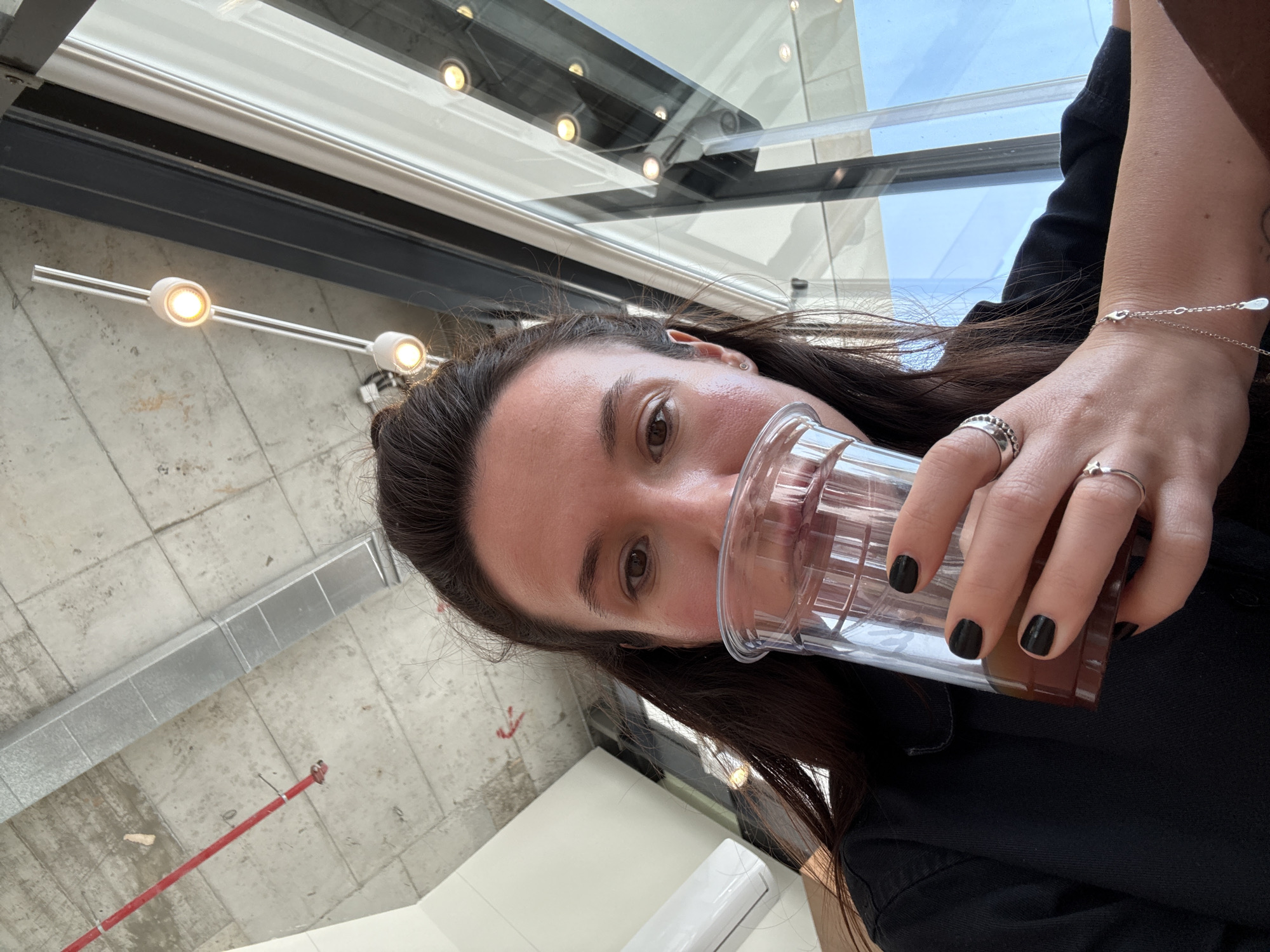 A woman is drinking from a clear plastic cup filled with a dark-colored beverage. She is holding the cup with her right hand, which has rings on multiple fingers and black nail polish. She is wearing a silver bracelet on her right wrist. The woman has dark hair and brown eyes, and she is wearing a black top. The image is taken from a low angle, looking up towards the woman. The background includes a modern building with a window. A sprinkler system is visible on the ceiling. The lights, glass, and ceiling suggest that the location may be a modern space in an urban area such as Tel Aviv, Israel. There is a white surface or object visible, possibly a table or a piece of furniture.