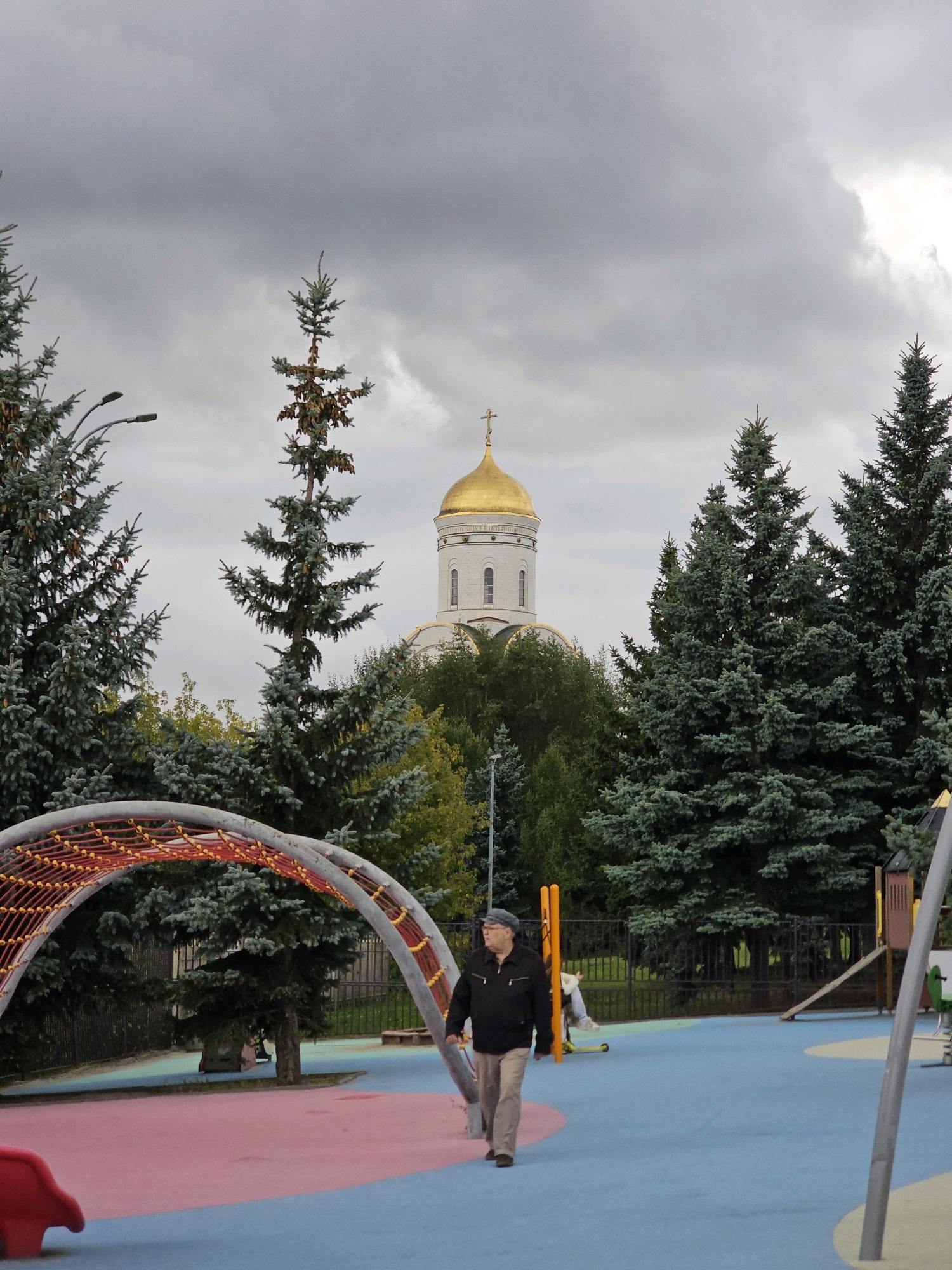 The image shows a man walking on a playground in Moscow, Russia. In the background, there is a church with a golden dome. The playground has a blue and pink surface, and a net structure. There are trees surrounding the playground, and a cloudy sky. The man is wearing a dark jacket and light-colored pants.