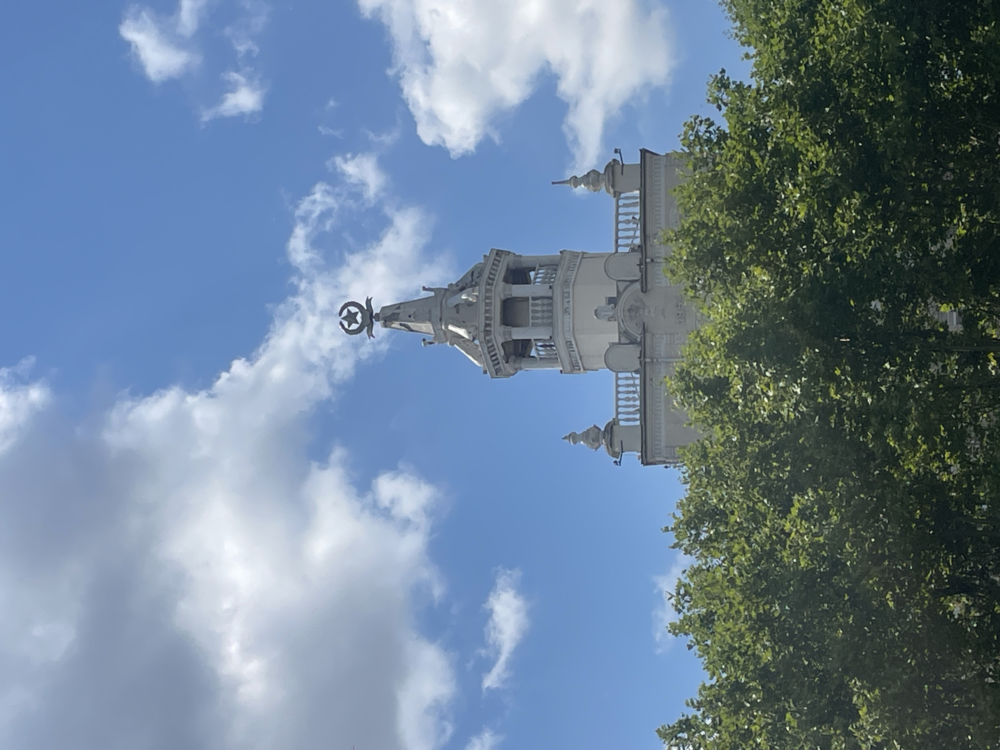 The image shows the upper portion of a white tower with a star on top, against a blue sky with white clouds. The tower has architectural details like columns, balustrades, and decorative spires. Below the tower, a dense green canopy of trees obscures the lower part of the building. The date "1954" is visible on the facade of the building beneath the trees. This building is the "House of the Officers" (Dom Ofitserov) located on Admiral Serebryakov Street in Novorossiysk, Russia.