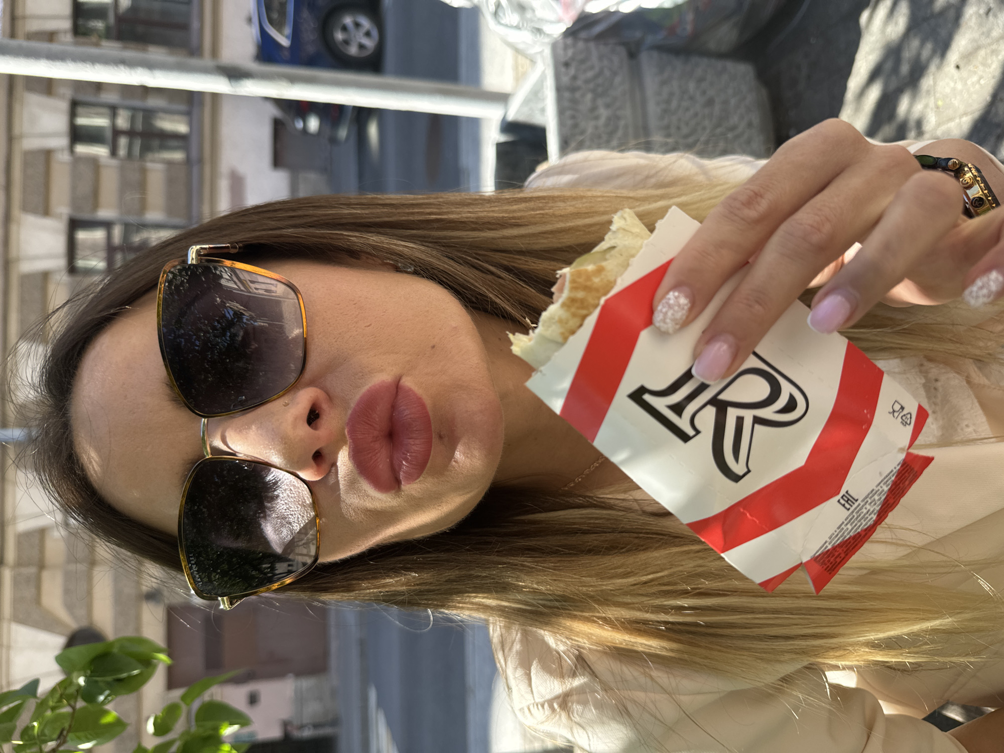 In this media file, a young woman is captured in a close-up shot, wearing sunglasses and holding a red and white wrapper with the letter "R" prominently displayed. Her lips are pursed, suggesting a playful or pouting expression. The background is blurred, but hints of a city scene with buildings and a vehicle can be discerned, consistent with the Saint Petersburg, Russia location context. The lighting suggests it is daytime, possibly late afternoon given the presence of shadows. The woman has long, blonde hair and her nails are painted with a decorative, possibly glittery, design. She is also wearing a bracelet. The wrapper she is holding appears to be for food, perhaps a snack or a wrap. The overall tone of the image is casual and perhaps a bit whimsical. Visible text includes the letter "R" on the wrapper and a small "EAC" logo.