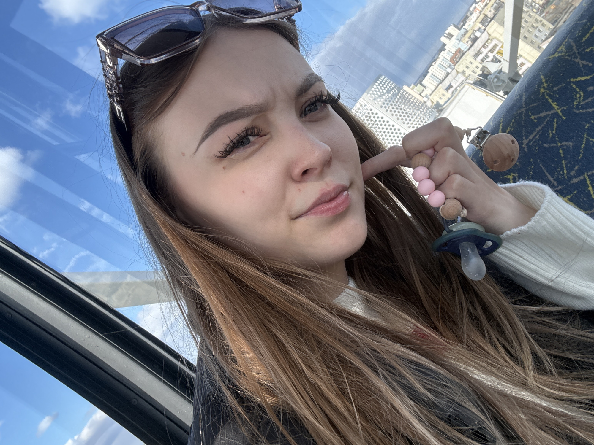 A young woman is taking a selfie indoors, possibly in a tall structure like a Ferris wheel or an observation tower given the urban background visible through the window. She has sunglasses perched on her head, long brown hair, and is touching her cheek with one finger. She is holding a baby pacifier on a beaded string. The window shows a partial view of the outside with buildings. It is unlikely that this image is from Yakutsk, Russia, as the architectural style and layout of the buildings are inconsistent with the typical cityscape of Yakutsk.