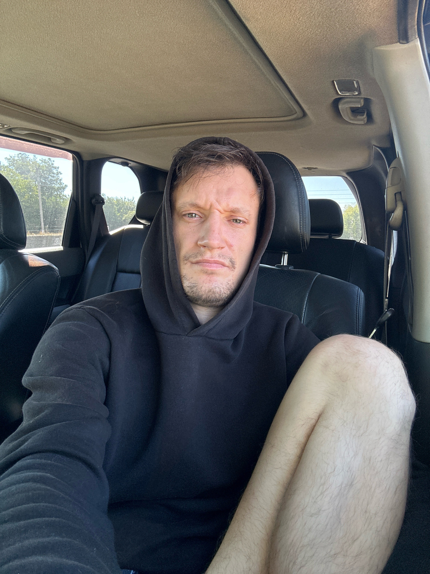 The image shows a man in the backseat of a car. He is wearing a black hooded sweatshirt and has his legs crossed. The interior of the car is visible, including the seats and the headliner. The man appears to be looking at the camera. The background outside the car windows shows trees and sky, suggesting an outdoor setting, possibly on the outskirts of Makiv, Ukraine.