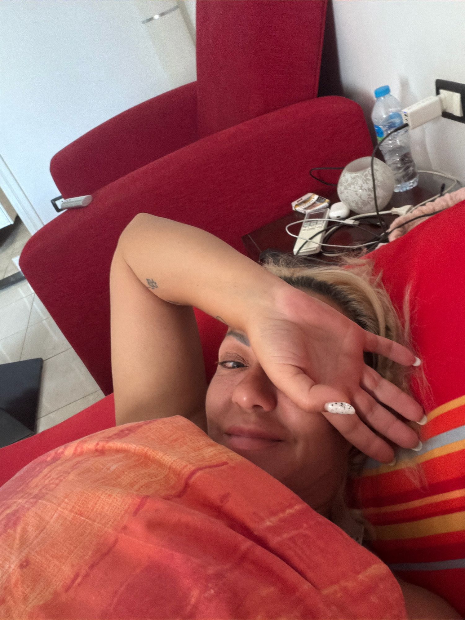 In an indoor setting, possibly a hotel room in Sharm El Sheikh, Egypt, a person is lying on a red sofa. The individual is partially covered by an orange and red patterned blanket. One arm is raised, covering a portion of their face, while the other arm is resting. Visible on the arm is a small tattoo. The person's hair is blonde and appears to be styled loosely. The background includes a white wall, a red armchair, and a bedside table with various objects such as a bottle of water, a decorative glass orb, electronic devices, and a remote control. A power adapter is plugged into the wall. The lighting suggests it is daytime.