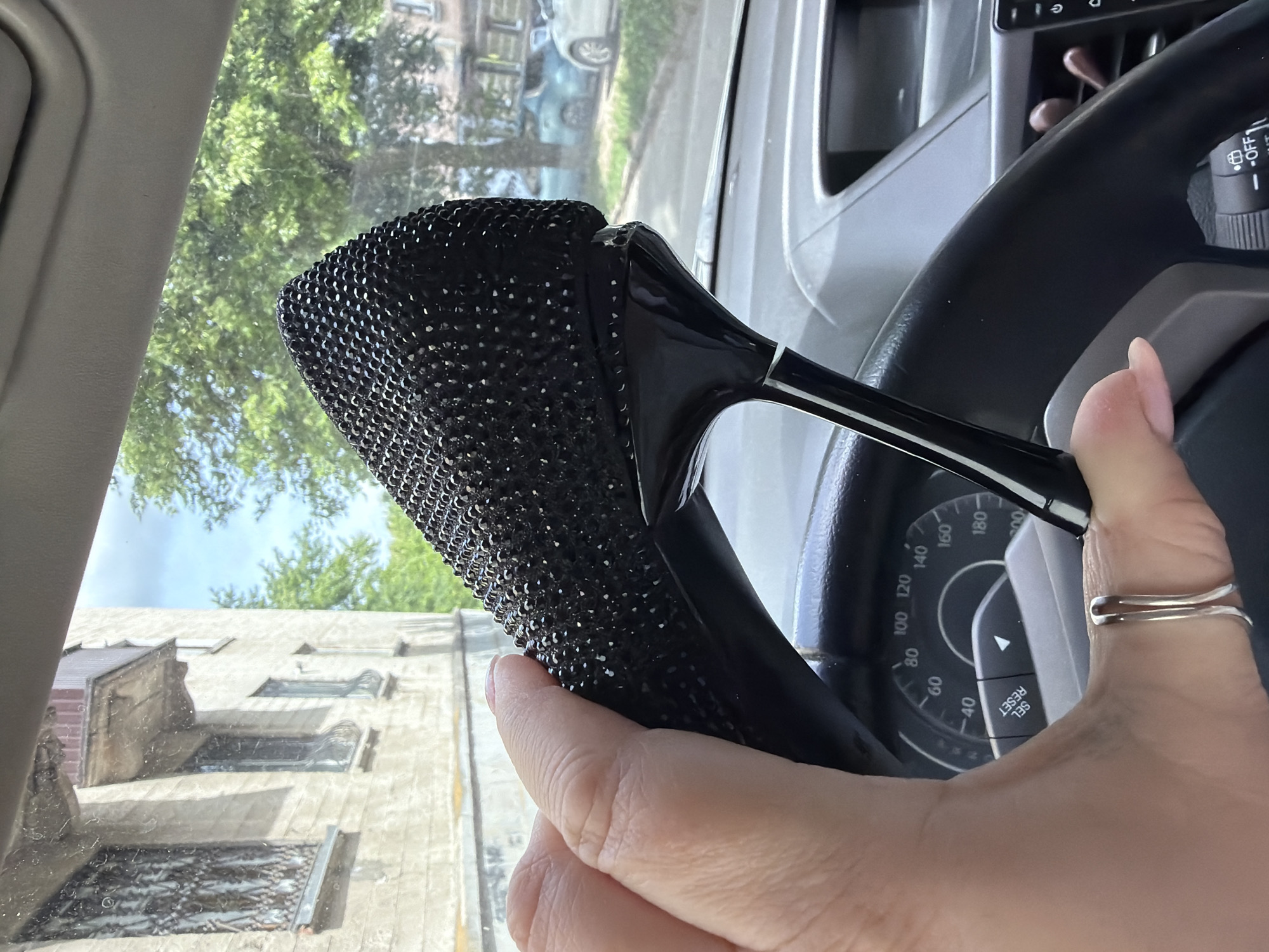 A right-hand view from inside a car shows a person holding a steering wheel. The steering wheel is black and has a decorative, possibly bejeweled, black cover. A hand, adorned with a silver ring on the ring finger, grips the steering wheel.
The dashboard and speedometer are visible behind the hand and steering wheel. Outside the car windows, there are trees and buildings with a clear sky above. The architecture of the buildings suggests a possibly European location. The specific location in Ternopil, Ukraine, cannot be confirmed from the given visual information alone.