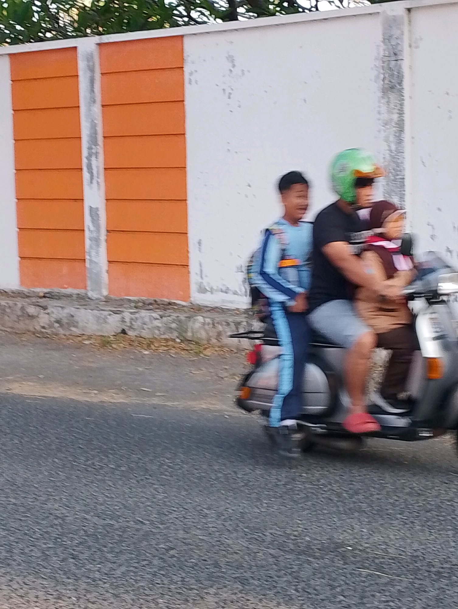 Three individuals are riding a scooter on a paved road in front of a wall with orange panels. The person in the front is wearing a green helmet and a black shirt. The person in the middle is a child wearing a blue and white tracksuit. The person in the back is wearing a maroon headscarf and a brown top. The scooter is gray and appears to be a vintage model. The wall behind them is white with vertical orange panels and a weathered texture. The road is asphalt, and there is some dry vegetation and debris on the side of the road. The scene suggests a typical day in Probolinggo, Indonesia, where scooters are a common mode of transportation.