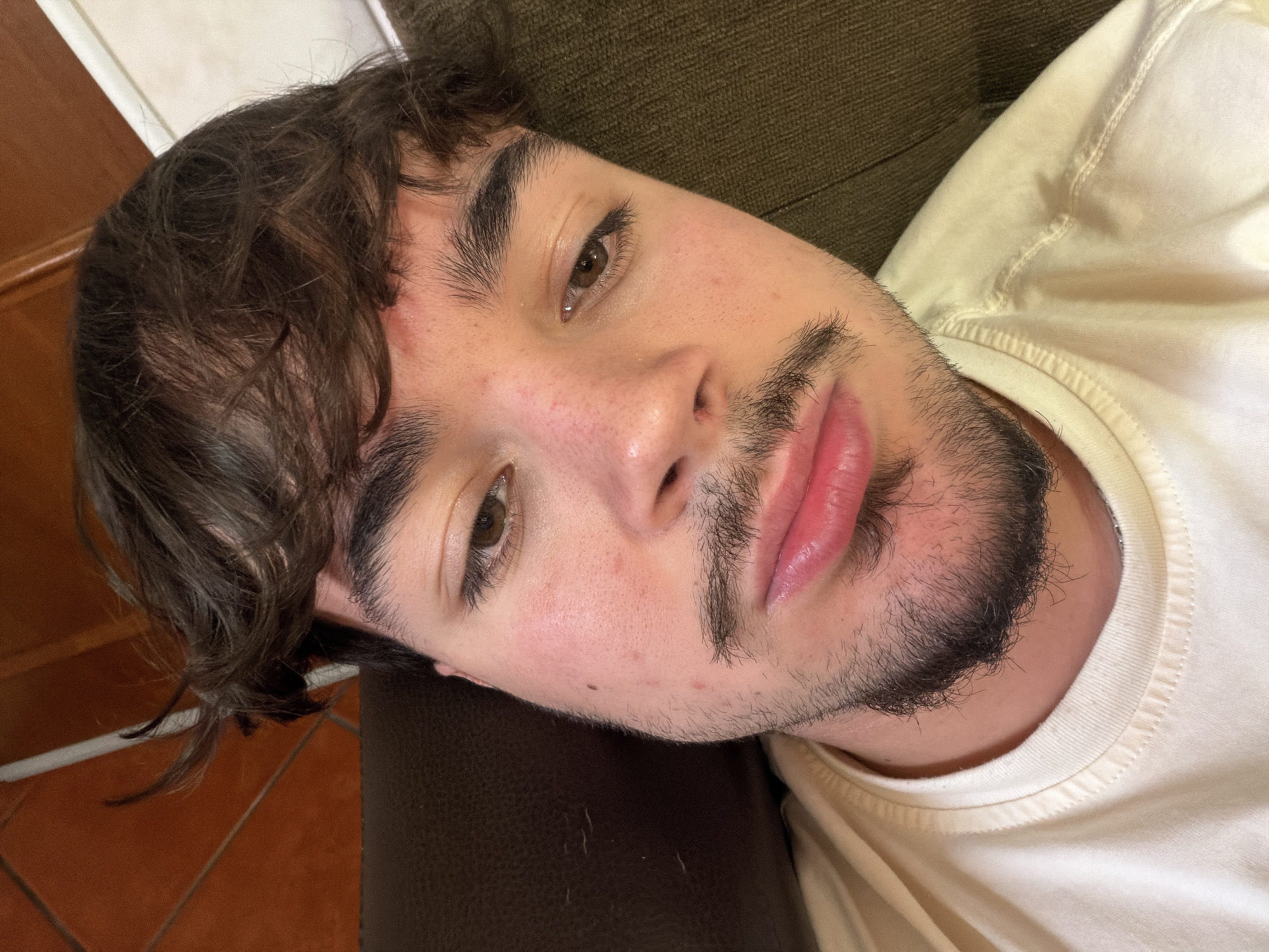 A close-up, angled shot of a young man lying down. He has dark brown hair with some waves, and a beard that's dense around the chin but sparse on the cheeks. His skin appears fair with a few blemishes, and his eyes are brown. He's wearing a light-colored t-shirt, and his head rests on a dark brown surface. Behind his head is an olive green, textured surface. The location in Vila Real, Portugal is not discernible from the image.