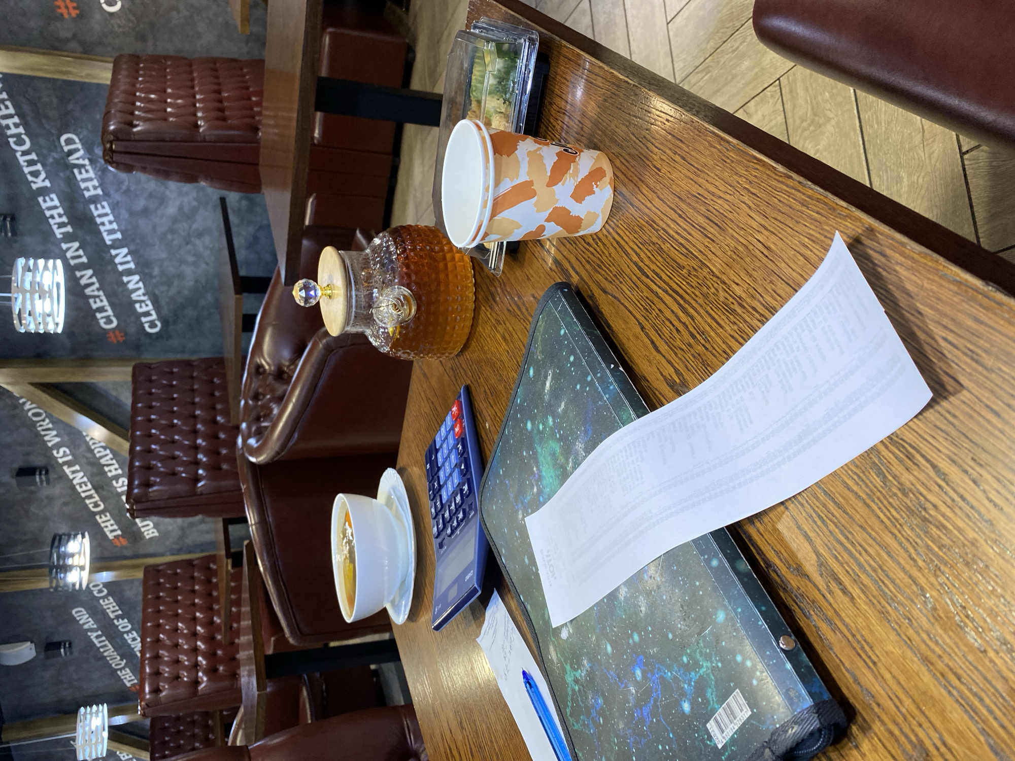 The image depicts a table in what appears to be a restaurant setting. On the table, there is a white bowl of soup, a calculator, a receipt, a pen, a patterned paper cup, a glass teapot filled with amber liquid, and a clear plastic container with food. The background shows booth seating made of dark red leather and a wall with motivational quotes written on it, including "#CLEAN IN THE KITCHEN" and "#CLEAN IN THE HEAD". The lighting suggests an indoor environment. The location is identified as the city of Ishimbay, Russia.