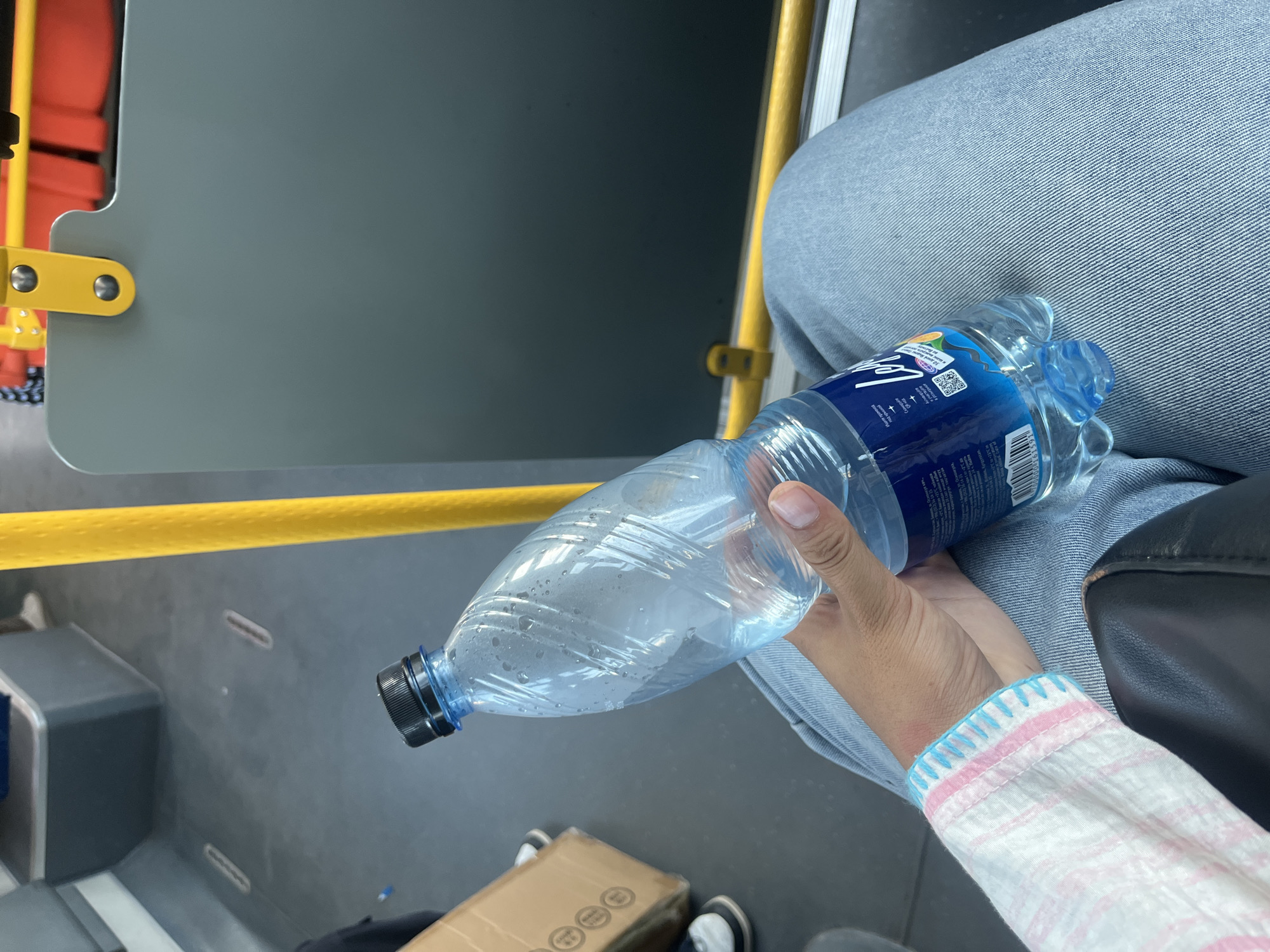 A person's hand holds a partially full plastic bottle of water with a black cap, situated inside what appears to be a public transport vehicle, possibly a bus, based on the yellow support pole and seats visible. The person is wearing a light-colored, checkered shirt with pink and blue trim and blue denim jeans. A cardboard box is visible in the background, to the left of the person. The floor of the vehicle is gray. The scene is an interior shot with ample lighting, suggesting daytime. There is no discernible indication of the location being specifically in Bishkek City, Kyrgyzstan.