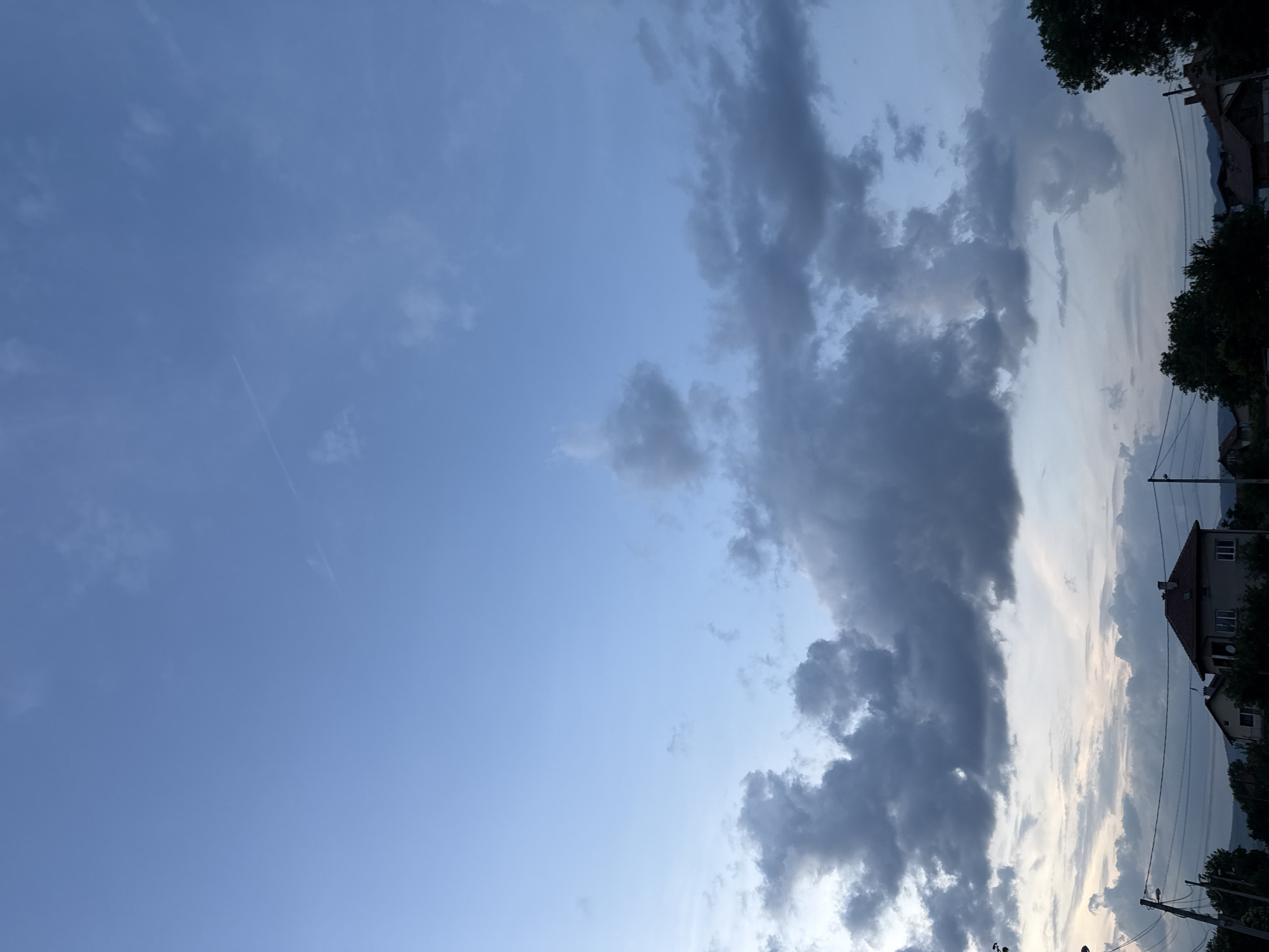 This image, taken in Mirovyane, Bulgaria, depicts an outdoor scene under a late evening or overcast sky. The upper sky displays a gradient from light azure blue at the zenith to a muted, paler blue towards the horizon. Two faint, linear airplane contrails are visible in the upper blue expanse, one longer and more distinct than the other. The lower half of the sky is dominated by a dense band of dark grey to near-black cumuliform clouds, with additional scattered, lighter grey cloud formations interspersed above. Below the cloud line, the upper portions of several residential structures with varying roof types are visible, notably a two-story building on the mid-right with a reddish-brown tiled roof and light-colored walls. The foreground features a dense line of dark green trees and bushes, partially obscuring the lower sections of the houses. On the left side, two wooden utility poles extend upwards, strung with multiple dark electrical or communication wires that crisscross horizontally across the mid-ground. No people, animals, or active vehicles are present in the frame.