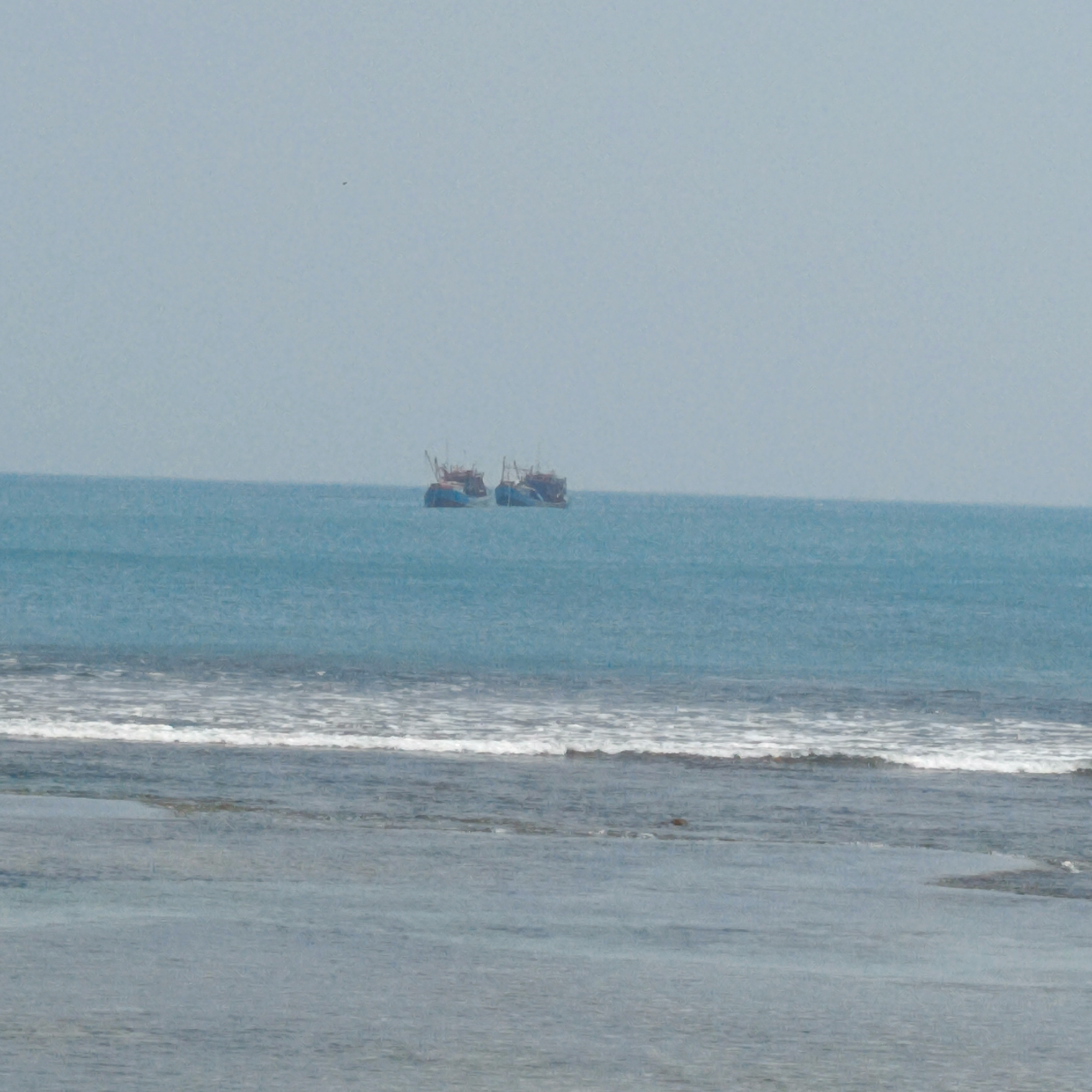 In the coastal waters off Pengubaian, Indonesia, two fishing boats are visible on the horizon. The boats, appearing to be of similar design, are anchored in the turquoise sea. The water in the foreground is calmer, with gentle waves lapping against a rocky shore, hinting at the shallow depth closer to land. The sky above is a pale, hazy blue, suggesting a partly cloudy or misty day. The overall scene is one of quiet activity at sea, characteristic of a fishing village in the region.