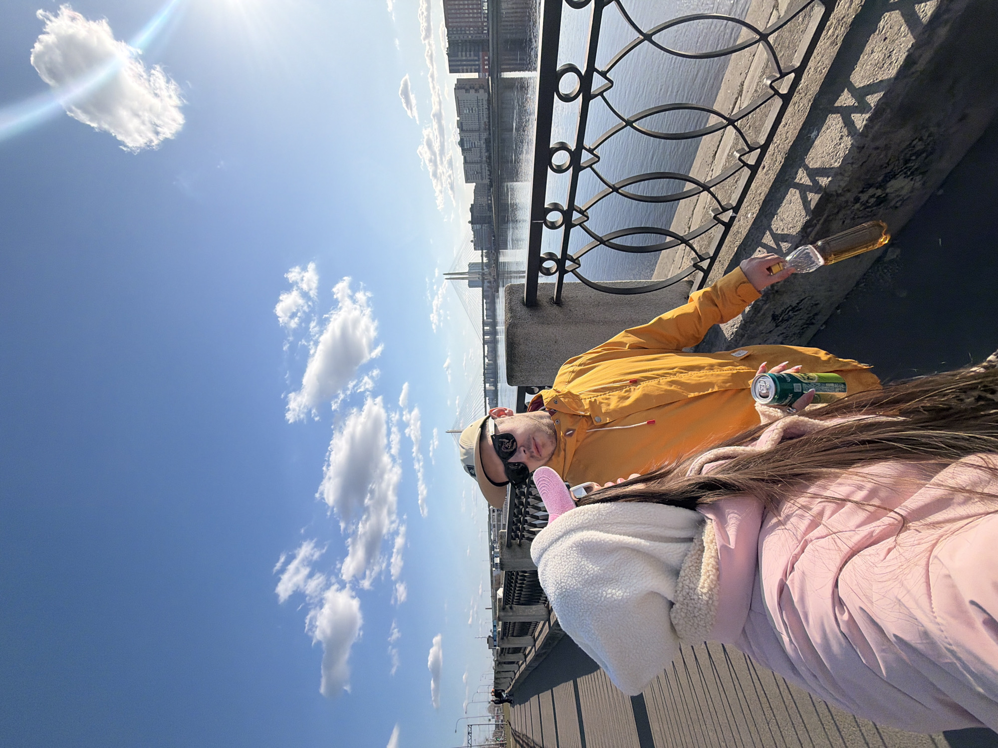 A selfie-style photograph captures two individuals on a sunny day along a paved embankment of the Neva River in Saint Petersburg, Russia.
The person taking the photograph (female, foreground left) is primarily visible from the back, wearing a light pink padded jacket with a white fleece hood and long dark hair. Her right hand holds a green aluminum beverage can, marked "Балтика 7" (Baltika 7 beer).
Next to her, a male subject (mid-ground) faces the camera, wearing a bright mustard yellow hooded jacket, dark sunglasses, and a beige baseball cap worn backward. He holds a clear plastic bottle containing an amber liquid in his left hand and a similar green beverage can in his right hand. Both individuals appear to be consuming beverages.
The background features the wide Neva River, with its calm surface reflecting the sky. Across the river, the prominent cable-stayed Bolshoy Obukhovsky Bridge with its two white pylons spans the waterway. Beyond the bridge, a line of multi-story residential or commercial buildings forms the distant city skyline.
The embankment consists of a concrete wall topped with an ornate black metal railing featuring a repeating pattern of connected oval shapes. The sky is bright blue with scattered white cumulus clouds, and strong sunlight from the upper right creates distinct shadows of the railing on the concrete wall and the subjects on the asphalt walkway.