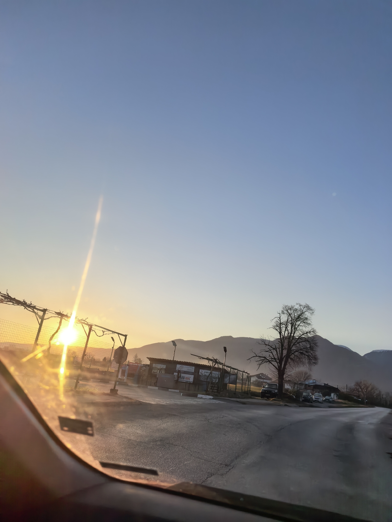 The image, taken from inside a vehicle, shows a sunrise over a rural or semi-rural landscape. The sun, low on the horizon, shines through a wire fence, creating a bright glare and lens flare. A small commercial building or structure is visible to the right of the fence, with some signage in a Cyrillic-based script. Beyond the building, a lone tree stands out against a backdrop of rolling hills or mountains. The road in the foreground is paved and appears damp, possibly from morning dew. The sky is a clear blue, transitioning to a lighter hue near the horizon. The scene suggests early morning on the outskirts of a town or village. The specific location within the city of Unknown, Unknown, is not discernible from the image.