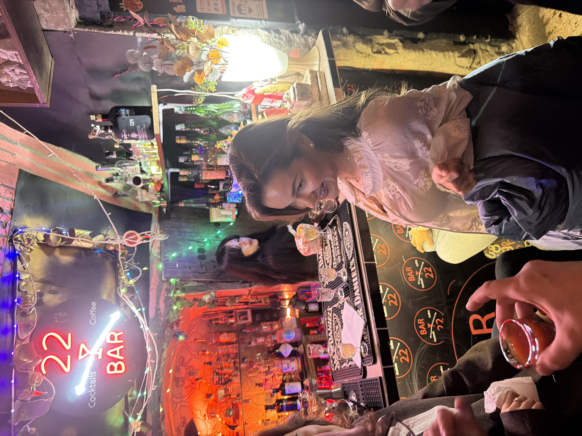The scene depicts the interior of "22 Bar" located at Akhvlediani Street 8 in Tbilisi, Georgia. The environment is dimly lit by various artificial light sources, including a prominent red neon sign displaying "22 BAR" with cocktail and coffee icons, and multicolored string lights draped across the ceiling.
Three individuals are visible. In the foreground on the left, a person's hands hold a shot glass containing a reddish liquid. This person is largely obscured, wearing a dark jacket, with a ring visible on the left hand. To their right, a female with long, dark brown hair and a light-colored, embroidered blouse smiles, looking towards the left side of the frame. Her left hand rests on a dark blue jacket. Behind the bar, a female with long dark hair, wearing a black top, stands facing left, positioned near various liquor bottles and a cocktail shaker. A fourth person's leg, clad in light green, is partially visible on the far right.
The bar counter features black "JACK DANIEL'S" rubber mats and a black facade with circular "22 BAR Cocktails Coffee" logos. Numerous bottles of alcohol are arranged on shelves behind the bar, illuminated by ambient lighting, predominantly red and green. Various pieces of glassware, including several shot glasses containing a pale yellow liquid, are present on the counter. The bar area also includes a tall, bright white lamp and an arrangement of dried flowers. The walls appear to be roughly textured or exposed, contributing to a rustic aesthetic.