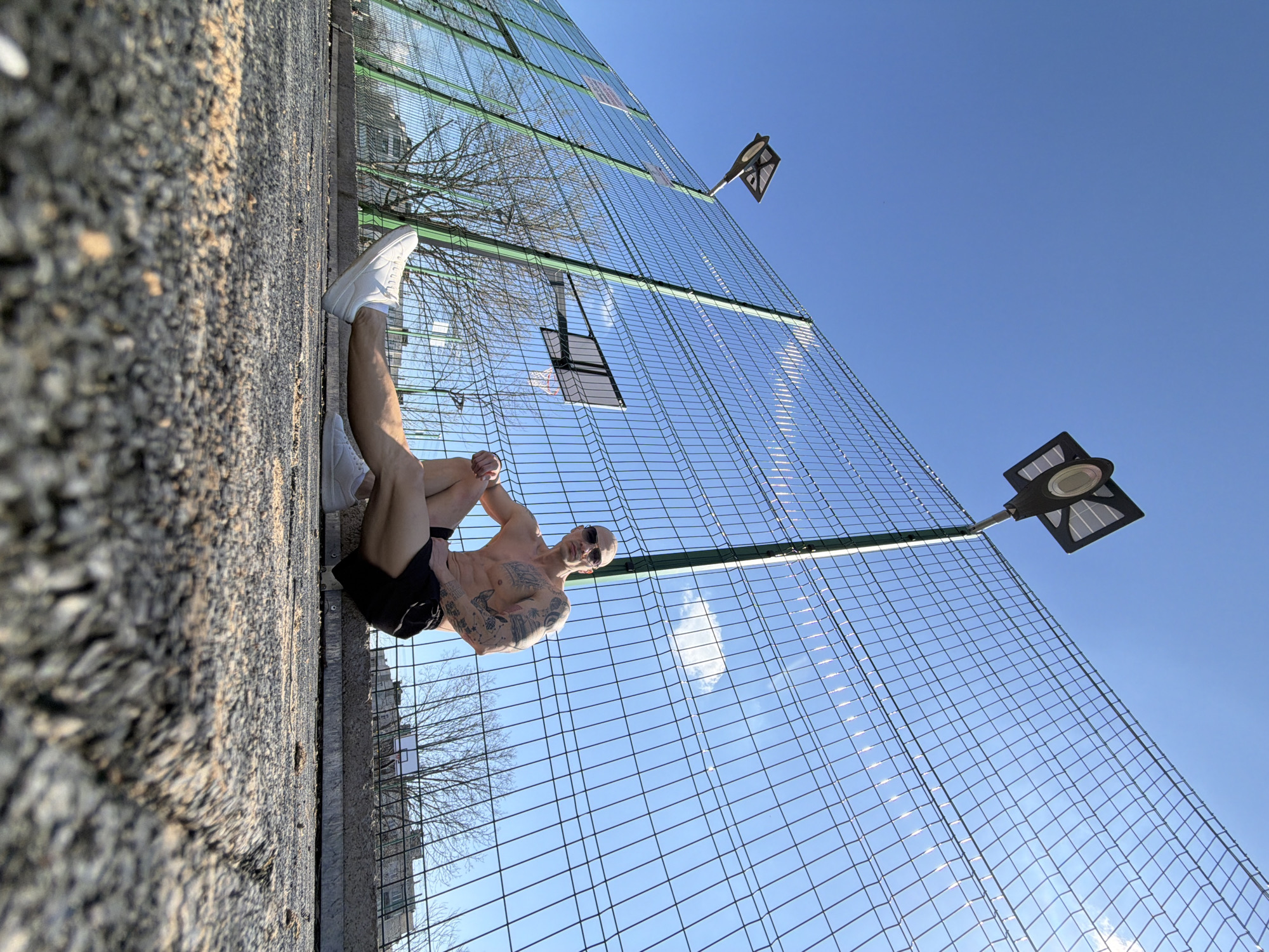 A low-angle shot depicts a fair-skinned man with a shaved head and visible tattoos on his right arm and torso, seated with his legs bent and crossed near a chain-link fence. He wears sunglasses, white sneakers, and black shorts. The fence, which forms the backdrop, is green-framed with two attached basketball hoops and two black, square solar-powered lamps mounted on poles. The surface on which the man is seated is asphalt. The sky is clear and blue. The location is most likely a recreational area in Pleven, Bulgaria.