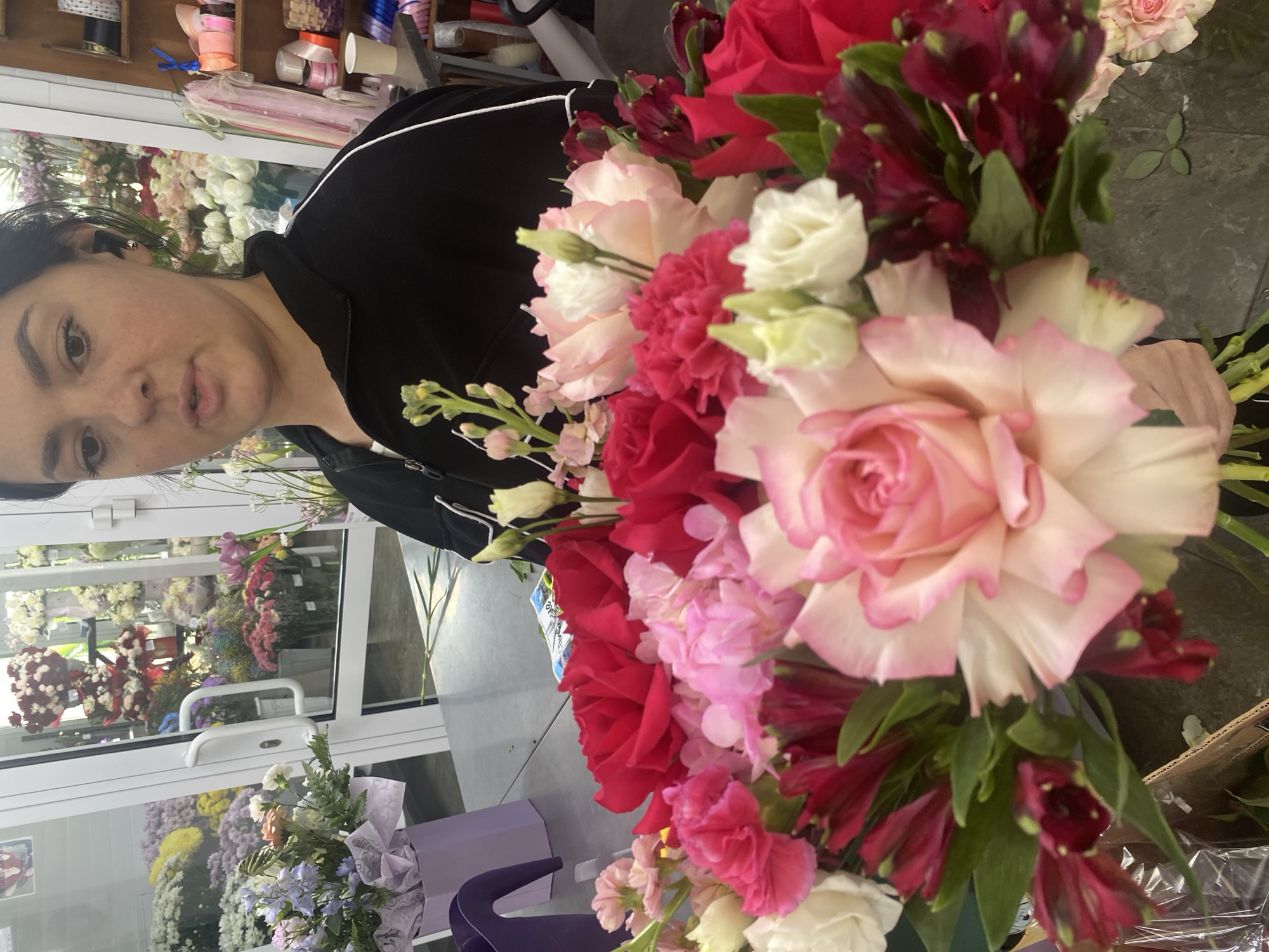 A woman in a black jacket is seen holding a bouquet of flowers. The bouquet contains pink roses, red roses, white lisianthus, pink carnations, and burgundy alstroemeria. The woman appears to be standing inside a flower shop. Through the glass doors of the shop, other bouquets of flowers can be seen. Shelves with rolls of ribbons are also visible. The location is likely a flower shop in Yekaterinburg, Russia.