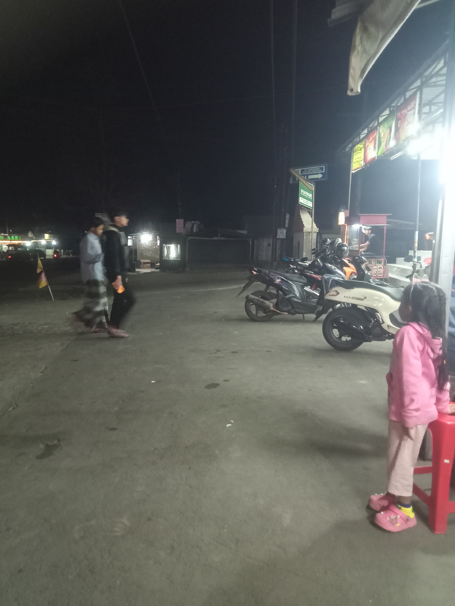 In the city of Urutsewu, Indonesia, at night, two men walk on a concrete pavement. One man wears a light blue shirt and a sarong, while the other wears a black shirt and black pants. In the background, several motorcycles are parked. To the right, a young girl in a pink hooded jacket and pink Croc-style shoes stands next to a red stool. She is facing away from the camera, looking towards the motorcycles. The scene appears to be in an outdoor market or roadside area, with some buildings and signs visible in the dim light.