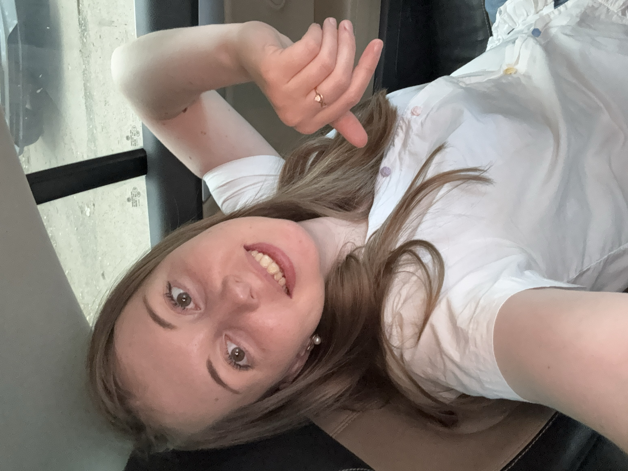 A young woman with long, light brown hair and brown eyes is seated in the back of a vehicle, taking a selfie. She is wearing a white t-shirt with colorful buttons. Her left arm is resting on the seat behind her, and her right arm is bent, with her hand positioned near her chin. She is smiling and looking at the camera. The background shows the interior of the car, including the headrest and window, with a blurry view of the outside.