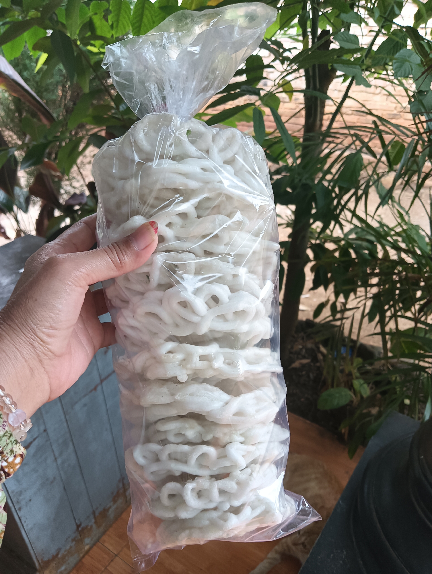 A close-up, high-angle shot captures a person's left hand holding a clear plastic bag filled with white, spiral-shaped crackers. The crackers are stacked vertically within the bag, and the plastic is gathered at the top with a twist tie. The background consists of lush green foliage, suggesting an outdoor or garden setting, possibly in Cluwak, Indonesia. The hand has a light brown complexion and is adorned with a bracelet made of beads. The overall impression is that of a casual, everyday moment, possibly a purchase or preparation of snacks.