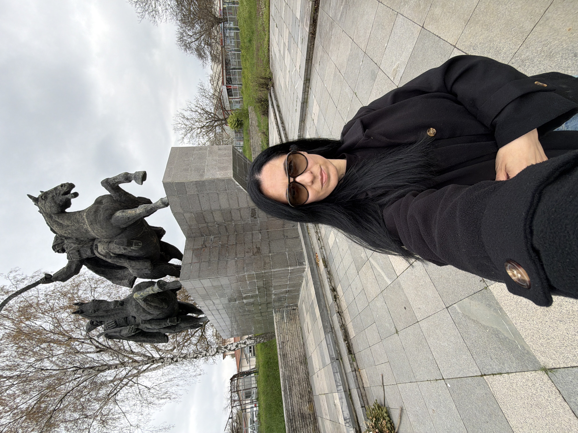 A woman with long dark hair, wearing sunglasses and a dark coat, is taking a selfie in front of the monument "Epic of 1877," located in Pleven, Bulgaria. The monument consists of a large stone structure supporting a bronze sculpture of horse riders. The ground is paved with square stones. Trees and buildings can be seen in the background. The sky is overcast.