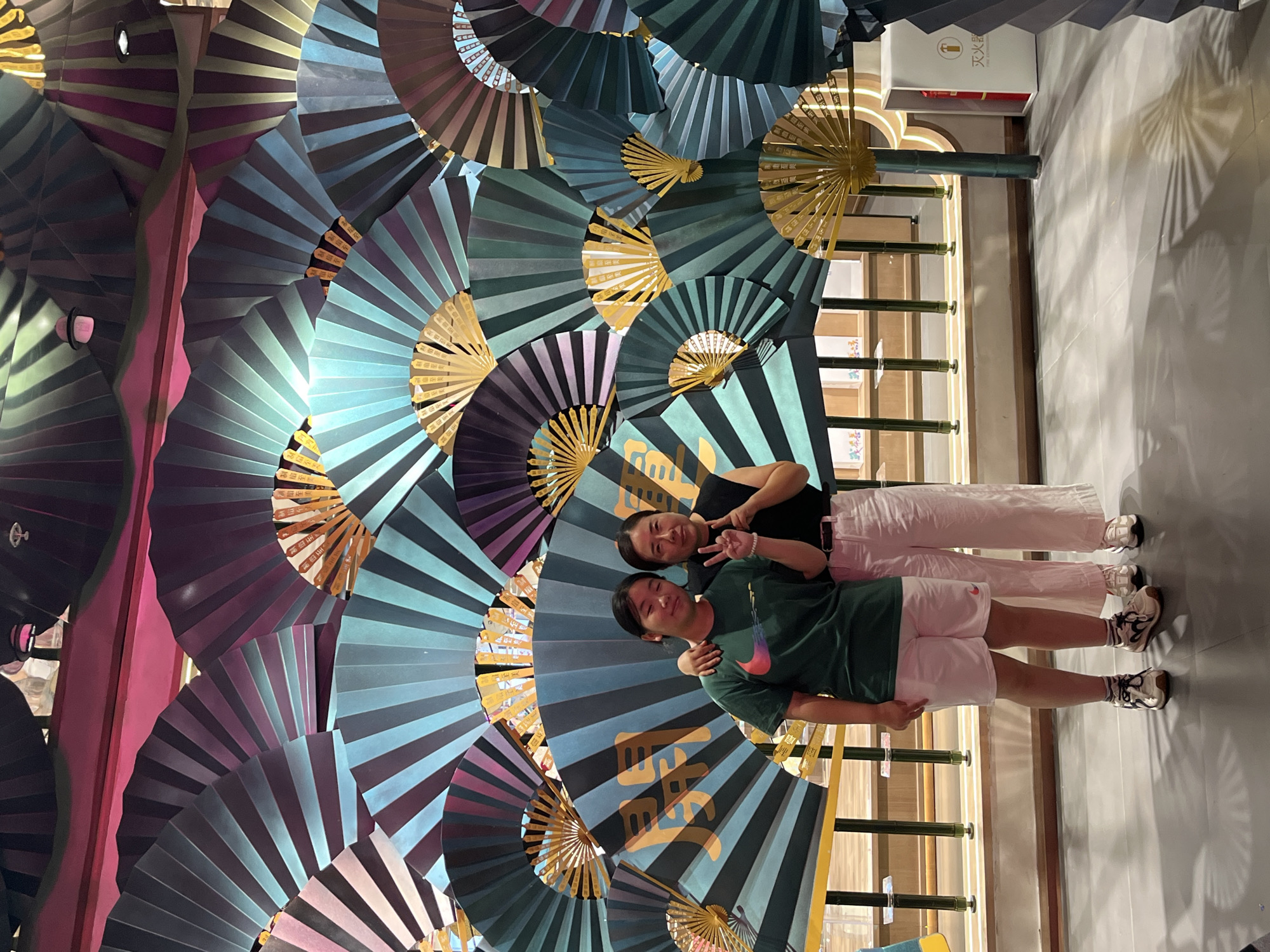 Two people, likely a mother and daughter, are posing for a photo in front of a decorative wall composed of numerous open paper fans in varying shades of blue, green, purple, and gold. The fans are arranged in a dense, overlapping pattern, creating a textured backdrop. Some of the gold fans have Chinese calligraphy written on them. The people are standing on a light gray tiled floor, and behind them, there is a light-colored wooden railing and vertical bamboo-like poles. To the right of the frame, a white fire extinguisher cabinet with Chinese text and a fire icon is visible. The lighting appears to be artificial, casting distinct shadows on the floor. The individuals are dressed casually: the person on the left wears a green t-shirt with a red and pink Nike logo and white shorts, while the person on the right wears a black t-shirt and loose-fitting light pink pants. The person on the right is making a peace sign with their fingers. The location is the Haizhu District, China, likely an indoor public space such as a mall or cultural center, suggested by the decorative elements and the presence of a fire extinguisher.
