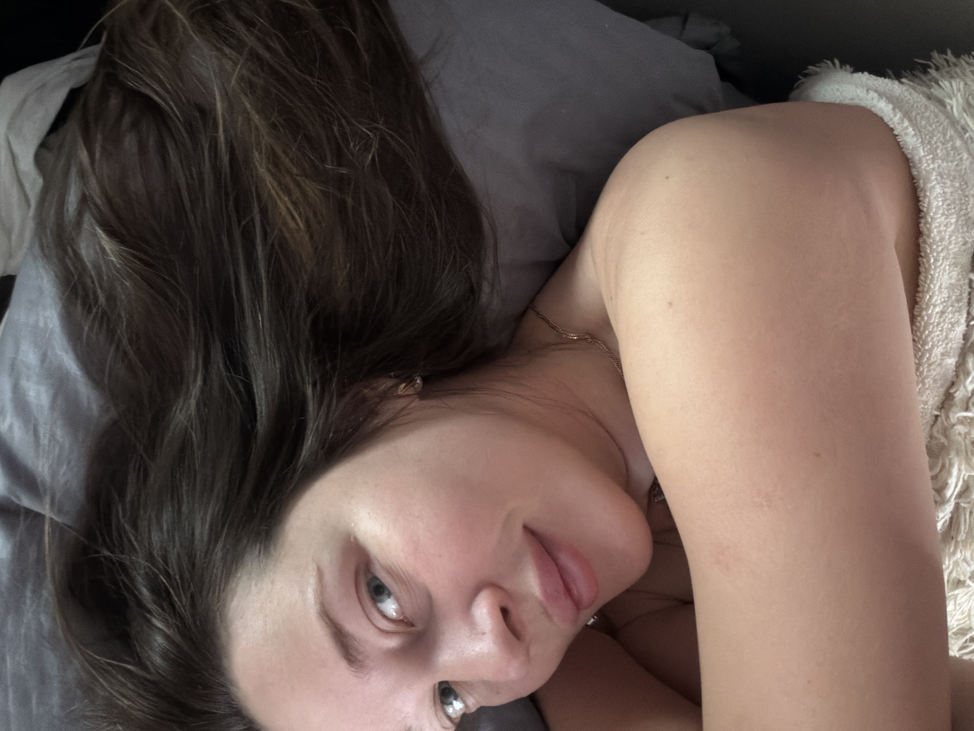 A woman with long brown hair is lying on a grey pillow. She is looking at the camera. Her skin appears to be fair. She is wearing a delicate gold necklace. A white fluffy towel is to her right. The location is not discernible from the image.